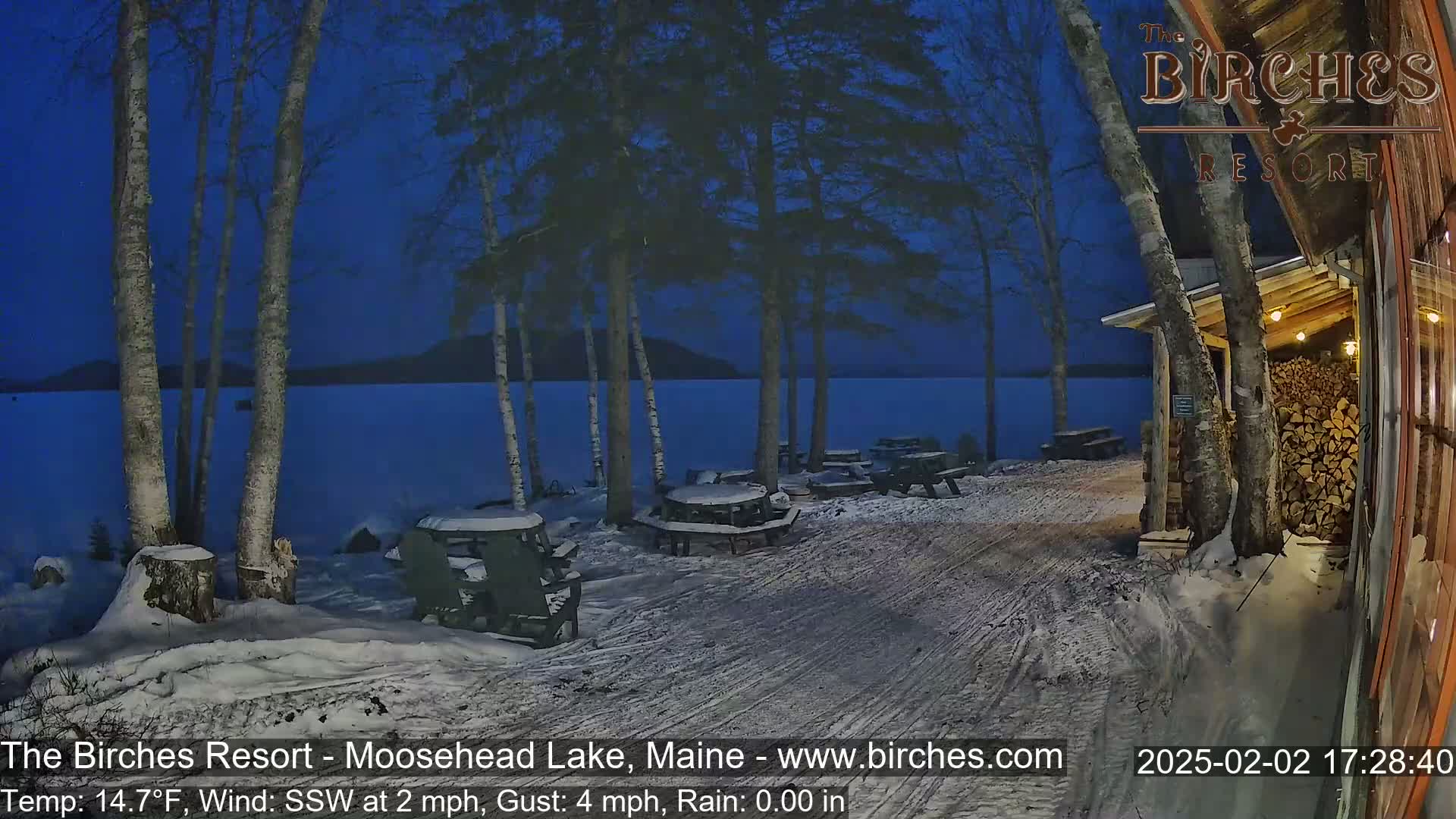Snow-covered picnic tables sit near a lake under a twilight sky, with a building and woodpile visible to the right.