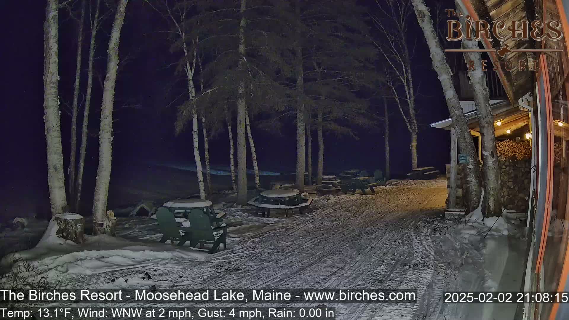 A snow-covered patio area with picnic tables and chairs sits outside a building at night under a dark sky.