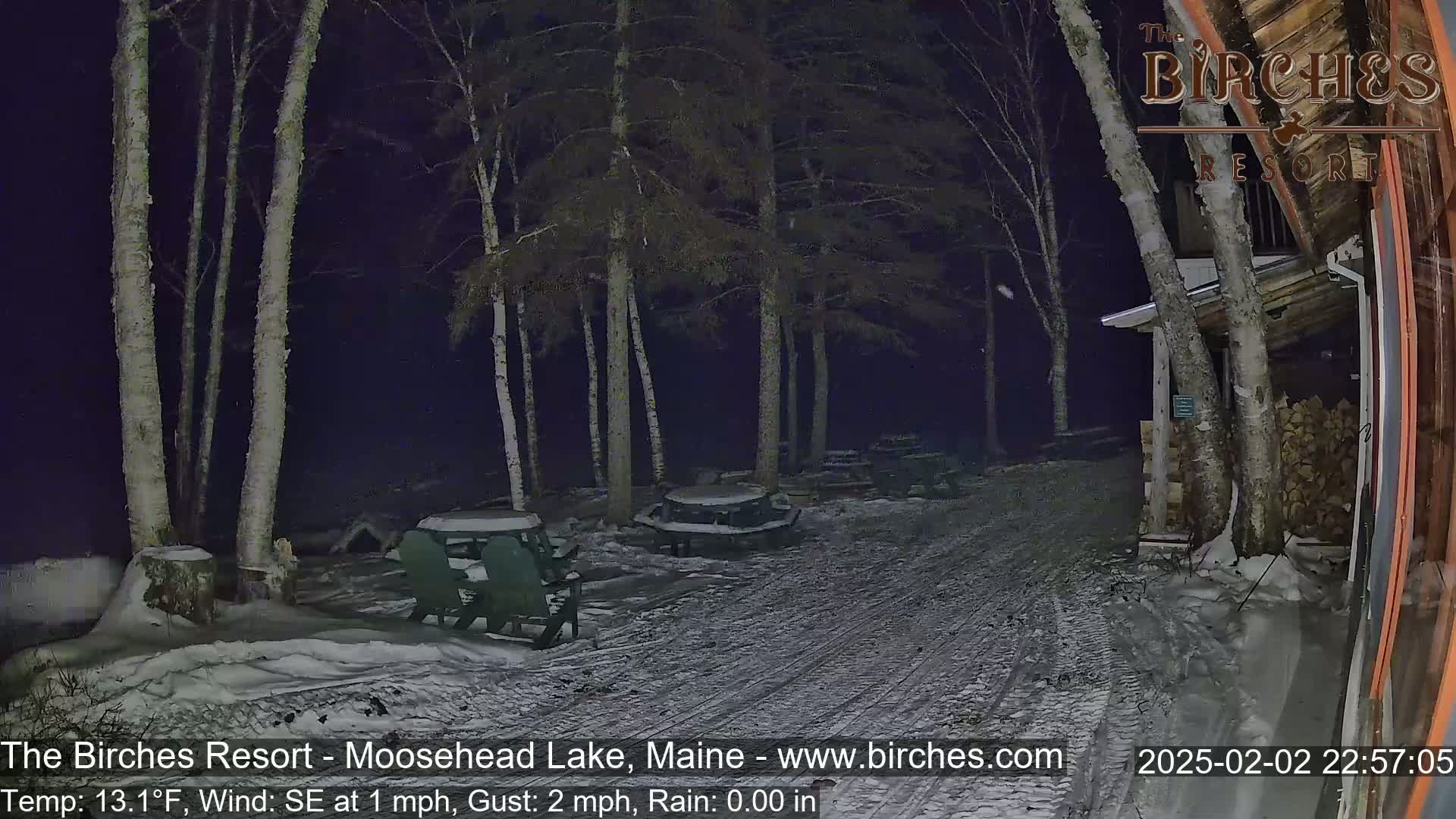 A snow-covered outdoor area at night, illuminated by artificial light, features several picnic tables and chairs under tall, slender trees.
