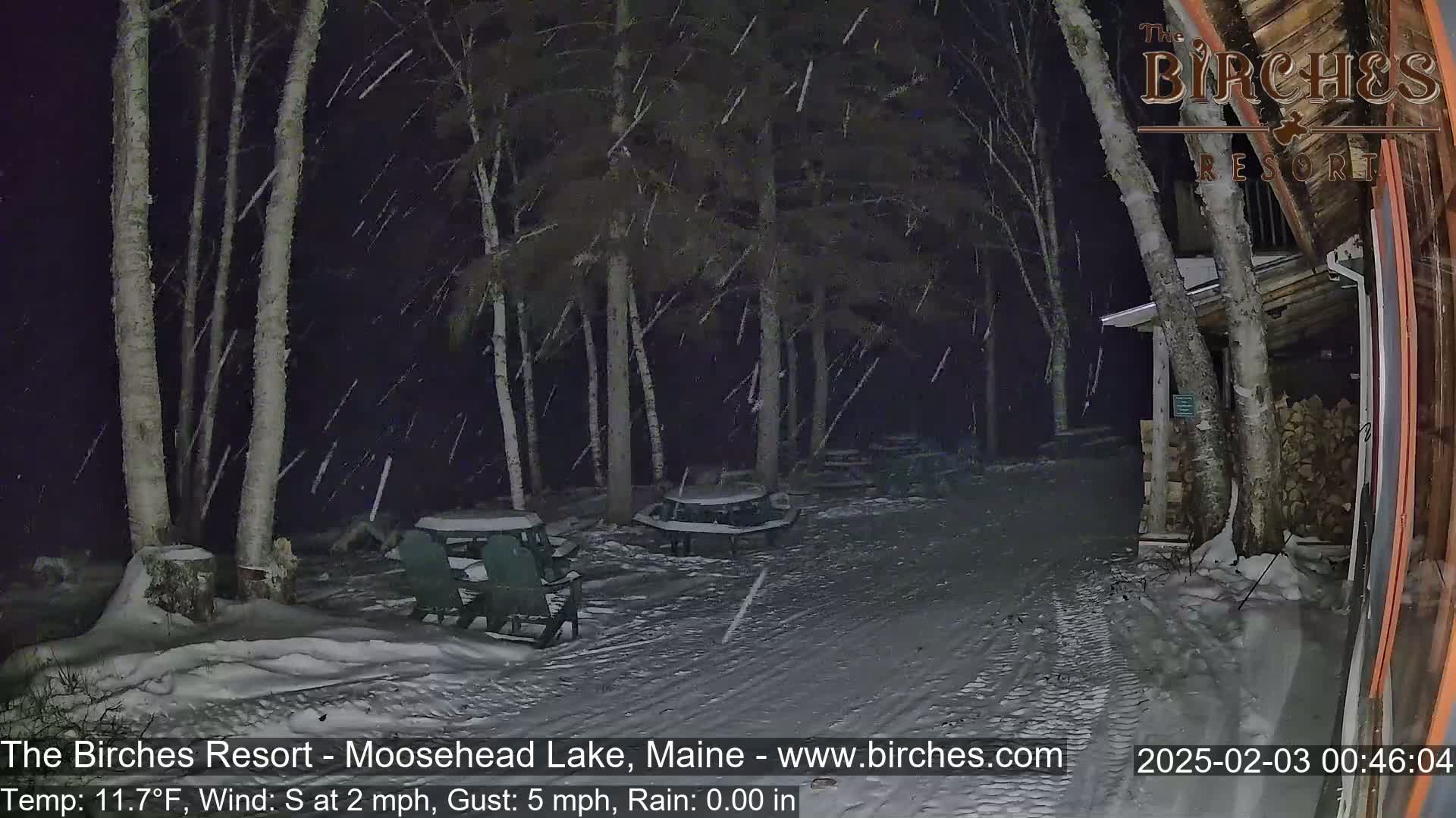 A snow-covered outdoor area with picnic tables and chairs is seen at night under falling snow near a building with stacked firewood.