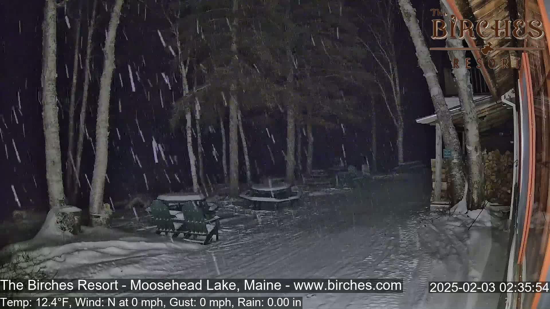 Snow falls at night on a snow-covered ground with picnic tables and Adirondack chairs near a building and trees.