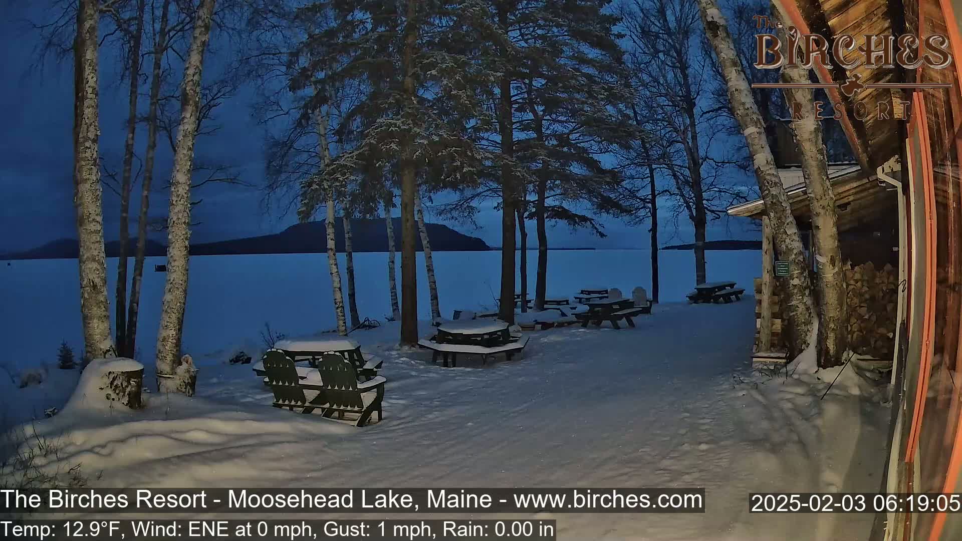 Snow-covered picnic tables sit outside near a lake under a twilight sky, with birch trees and a wooden building visible.