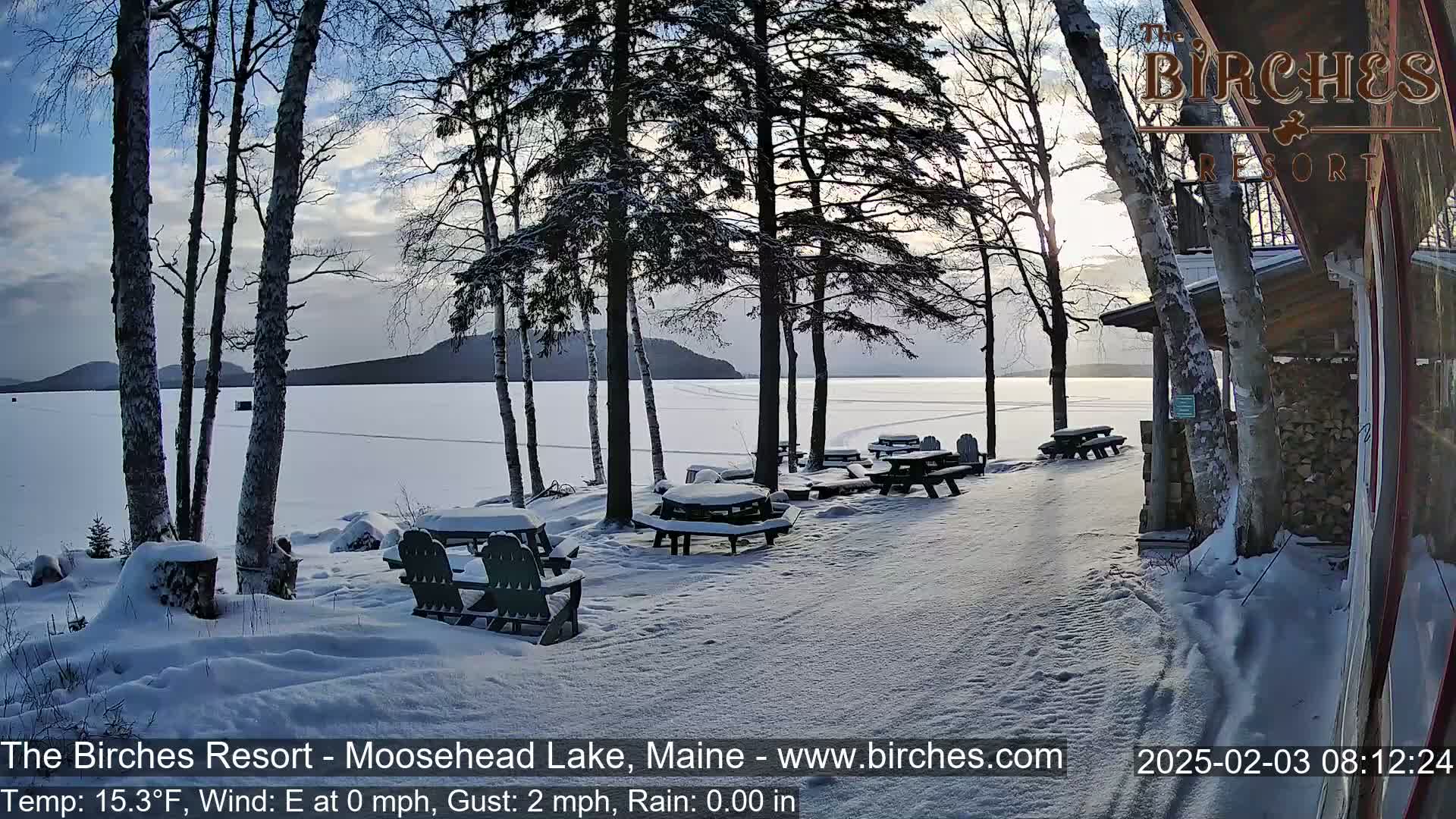 A snow-covered lakeside area with picnic tables and chairs, under a partly cloudy sky.