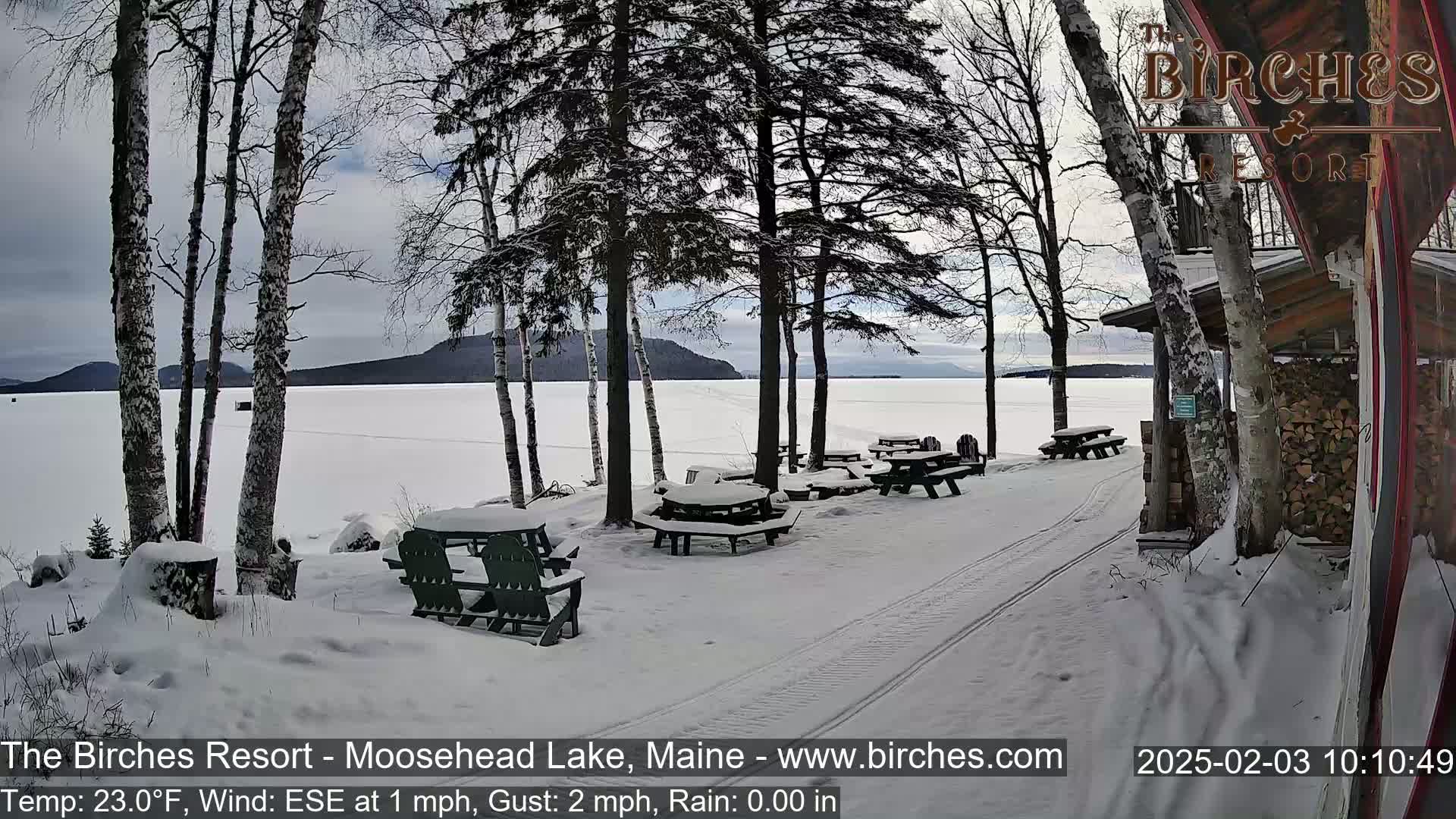 Snow-covered picnic tables and chairs sit beside a frozen lake under a cloudy sky.