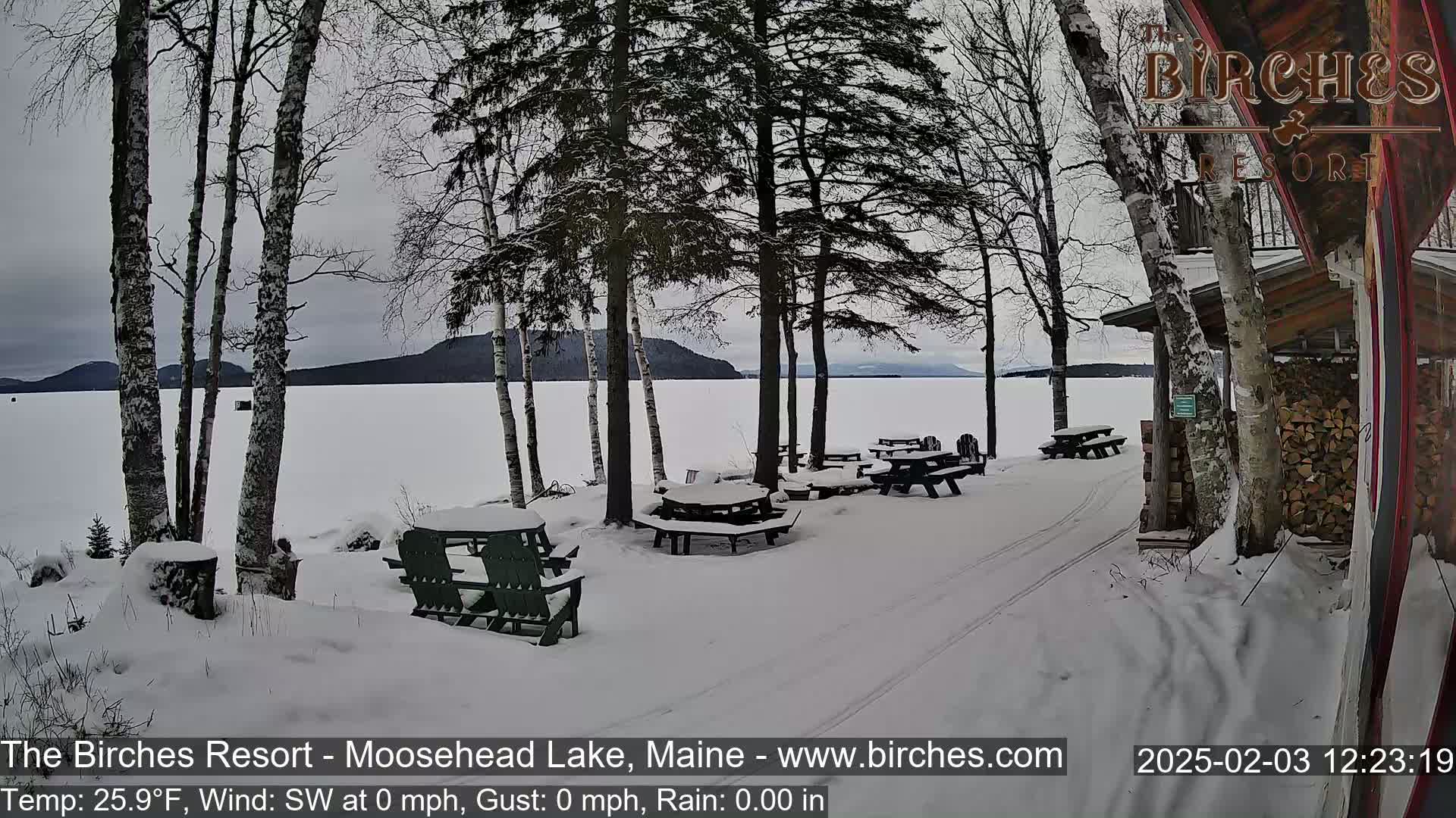 A snow-covered lakeside picnic area with several empty tables and chairs sits near a building, under a cloudy sky.