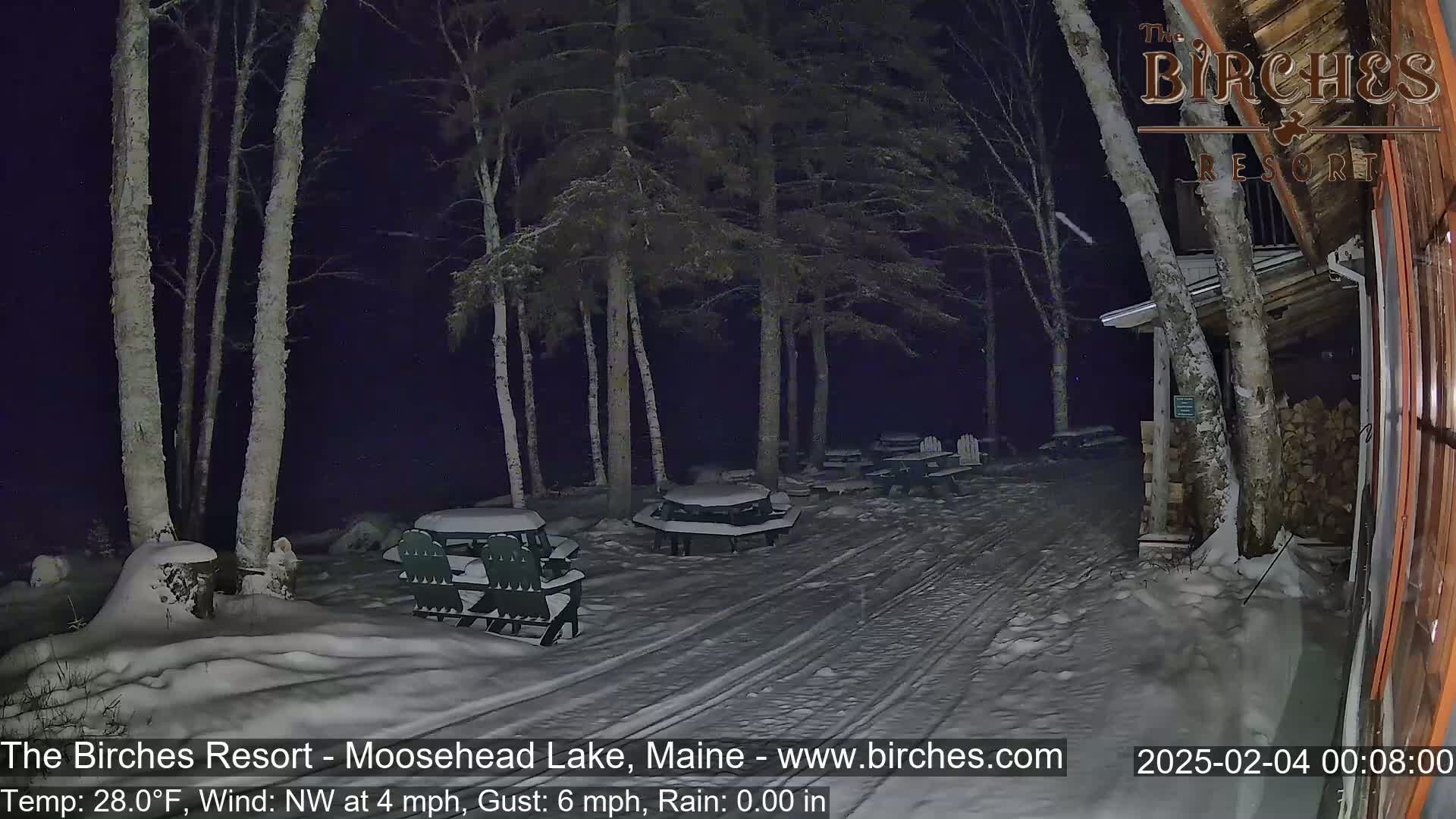 A snow-covered patio area with several picnic tables and chairs is visible at night, under a dark sky with a northwest wind.