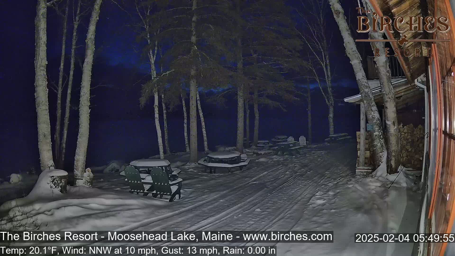 A snow-covered outdoor area at night, illuminated by artificial light, shows several picnic tables and chairs under a dark sky with a NNW wind at 10 mph and gusts up to 13 mph.