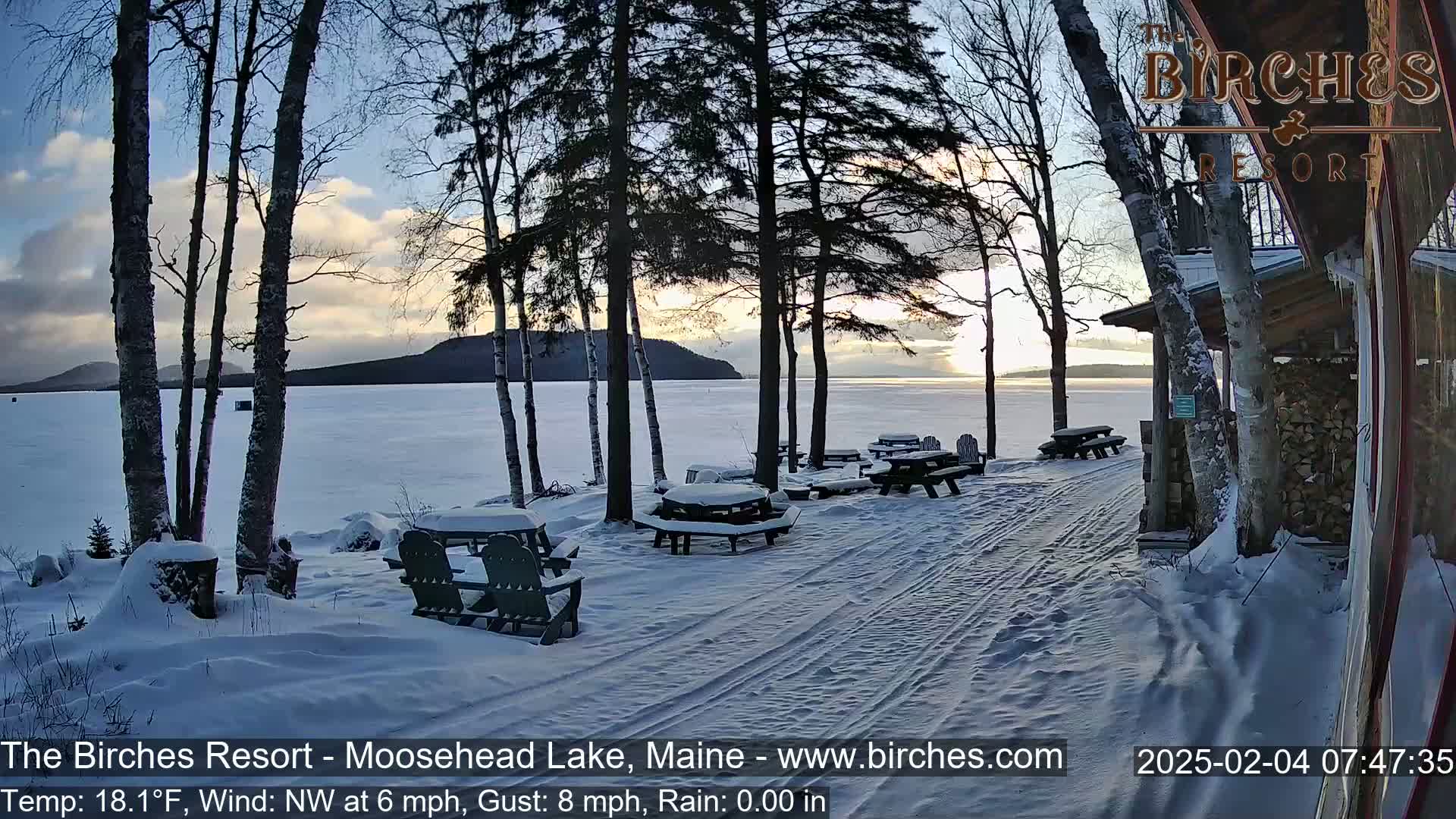 Snow-covered picnic tables sit near a snow-covered shoreline by a frozen lake at sunrise, with several birch trees in the foreground.