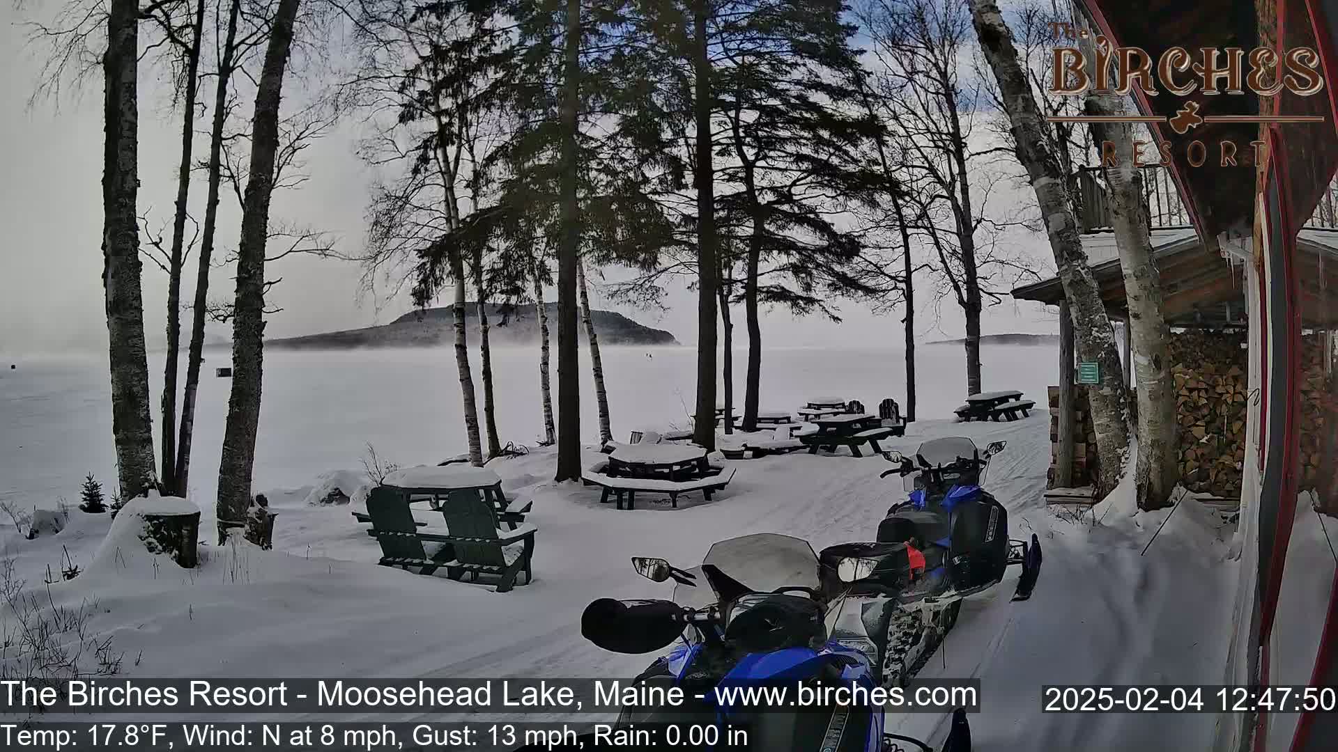 Snow-covered picnic tables and snowmobiles are parked near a lakefront building on a foggy, snowy day with a north wind.