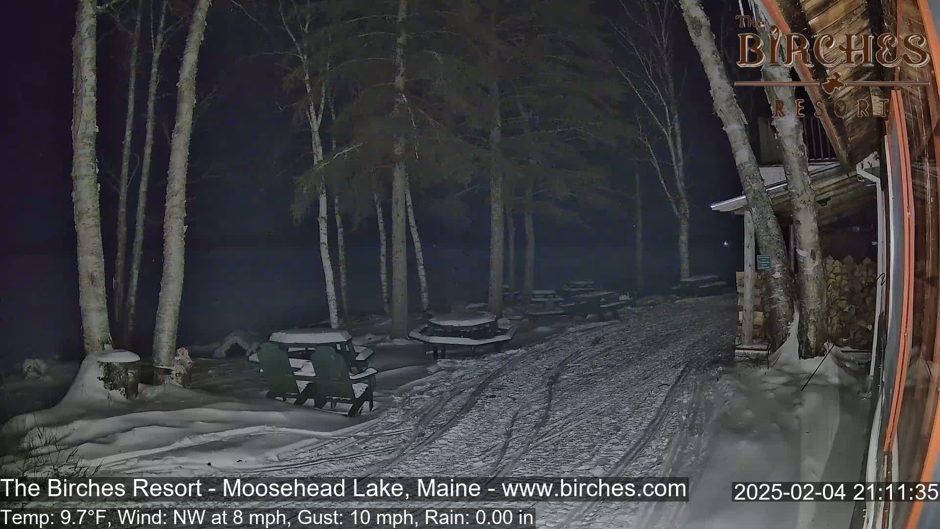A snow-covered outdoor area at night, with picnic tables and a building, under a dark sky with a northwest wind.