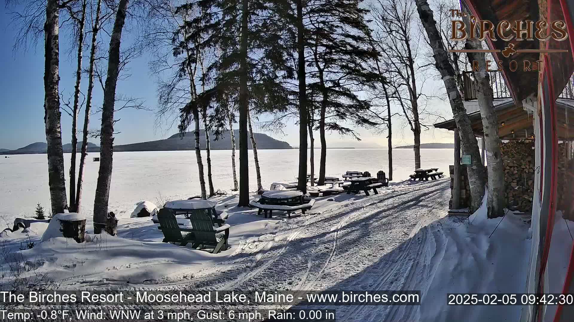 A snow-covered lakeside patio with picnic tables and Adirondack chairs overlooks a frozen lake under a sunny sky.