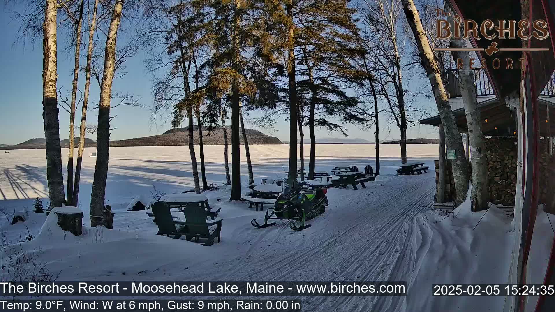 A snow-covered lakeside scene features a snowmobile parked near picnic tables and a resort building under a clear, sunny sky.