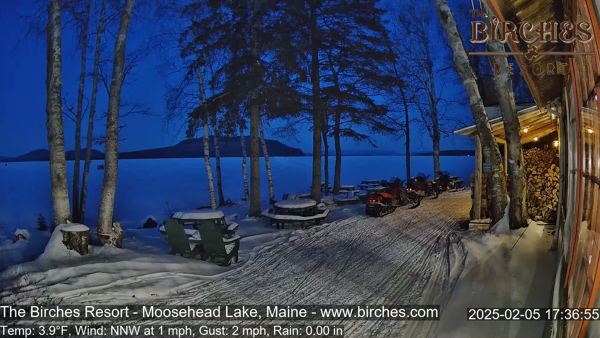 Snow-covered picnic tables and snowmobiles sit near a lakeside resort under a twilight sky, with a light wind.