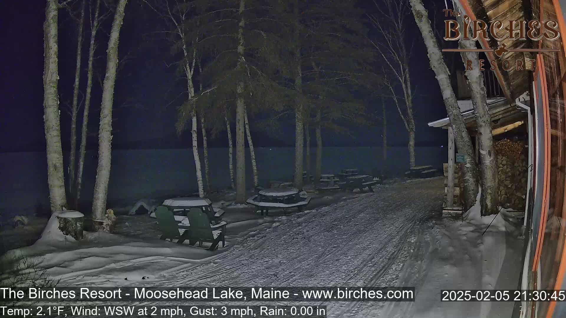A snow-covered path leads to a lakefront resort at night, with several snow-covered picnic tables and chairs situated near a building under a dark, clear sky.