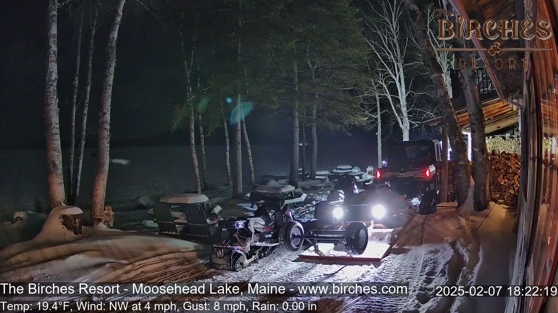 At night in snowy conditions, several snowmobiles are parked near a building with a woodpile, and one is being towed on a trailer by a utility vehicle.