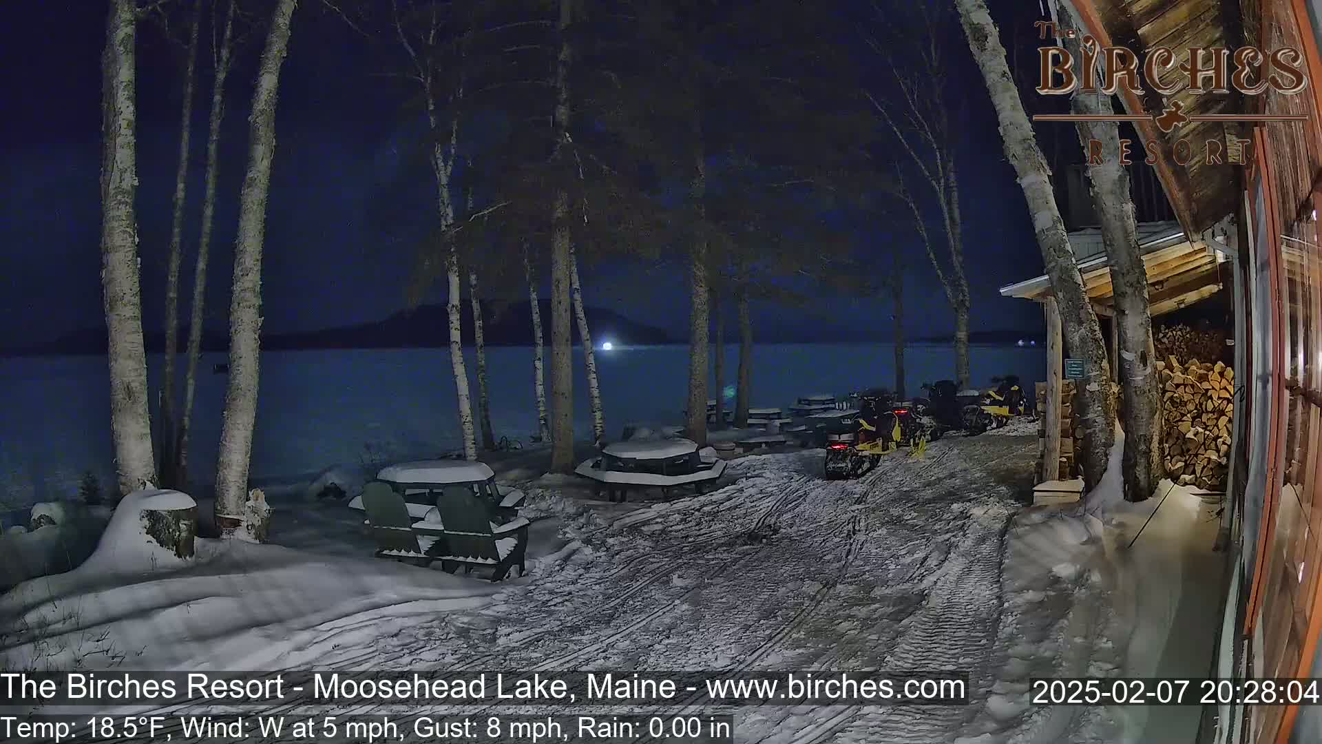 A snow-covered lakeside area at night, with several snowmobiles parked near a building and picnic tables under birch trees, with a calm lake and dark sky in the background.
