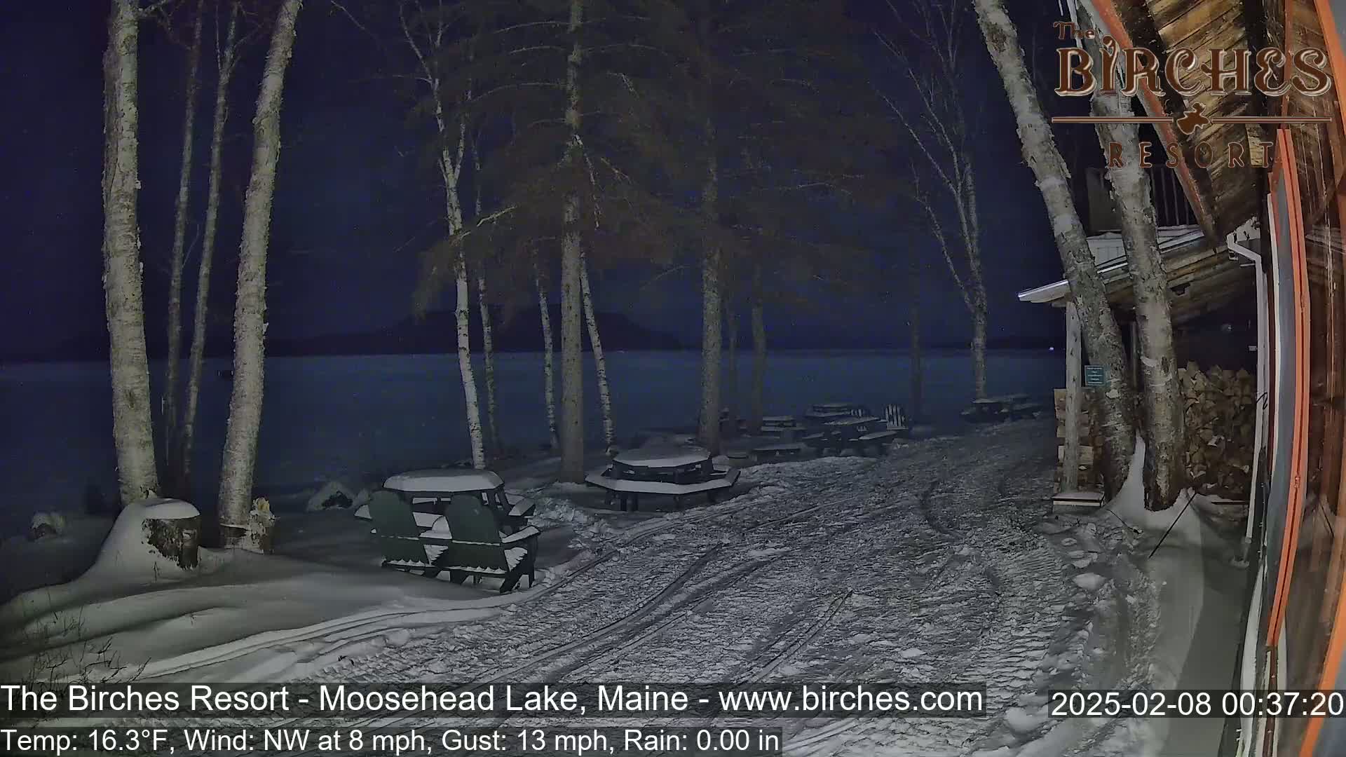 A snow-covered lakeside picnic area at night, with several empty tables and chairs, under a dark sky and a northwest wind.