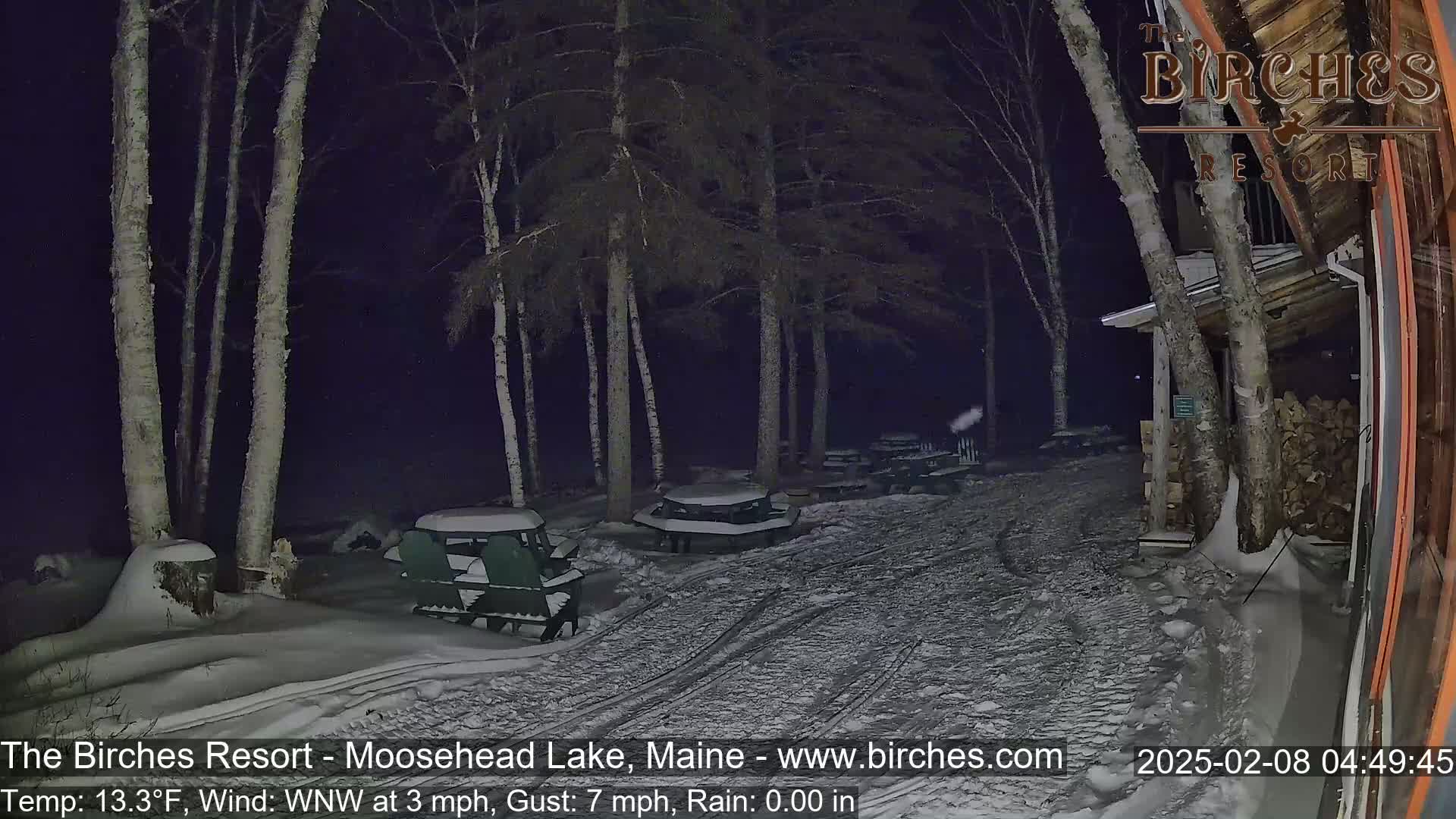 A snow-covered outdoor area at night with picnic tables and Adirondack chairs near a building under a dark sky.