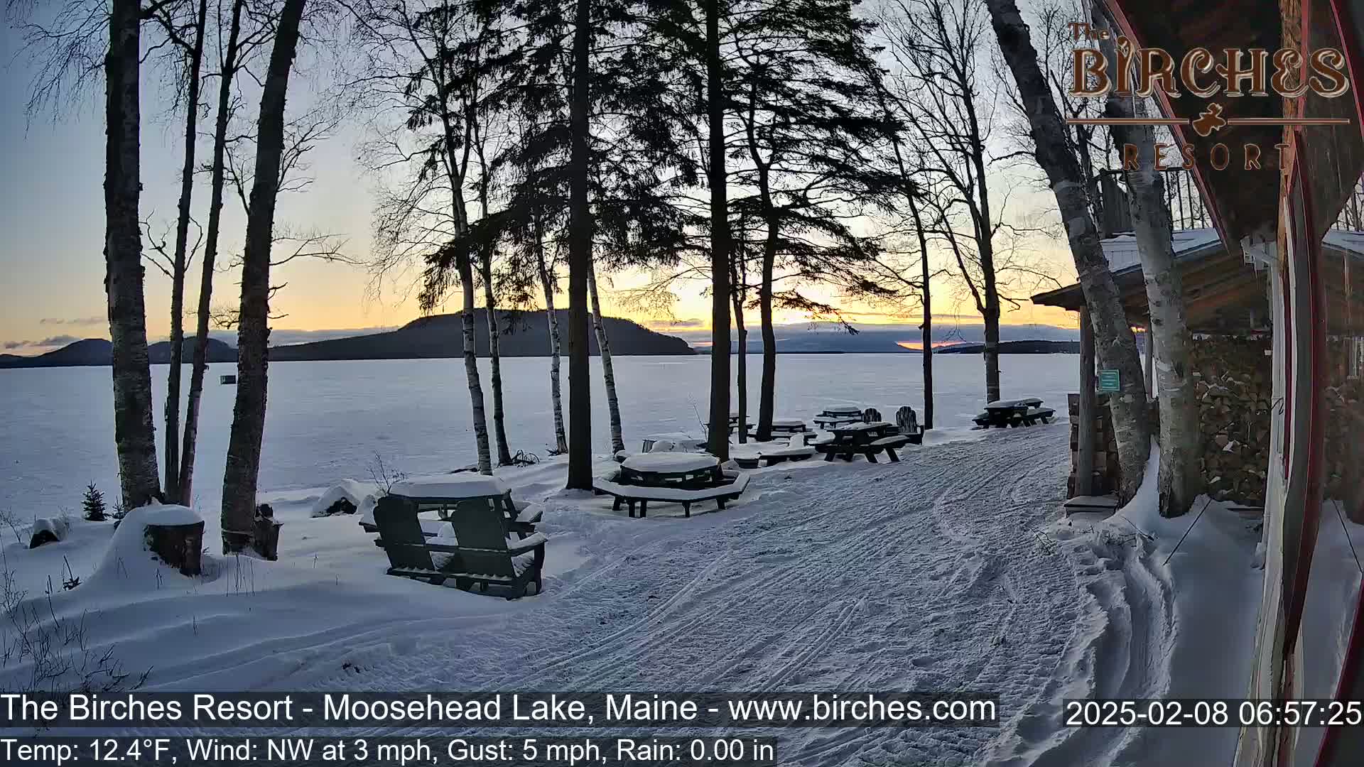 Snow-covered picnic tables and Adirondack chairs sit near a lake at sunset, with a wooden building in the background and bare trees in the foreground; the weather is cold with a light northwest wind.