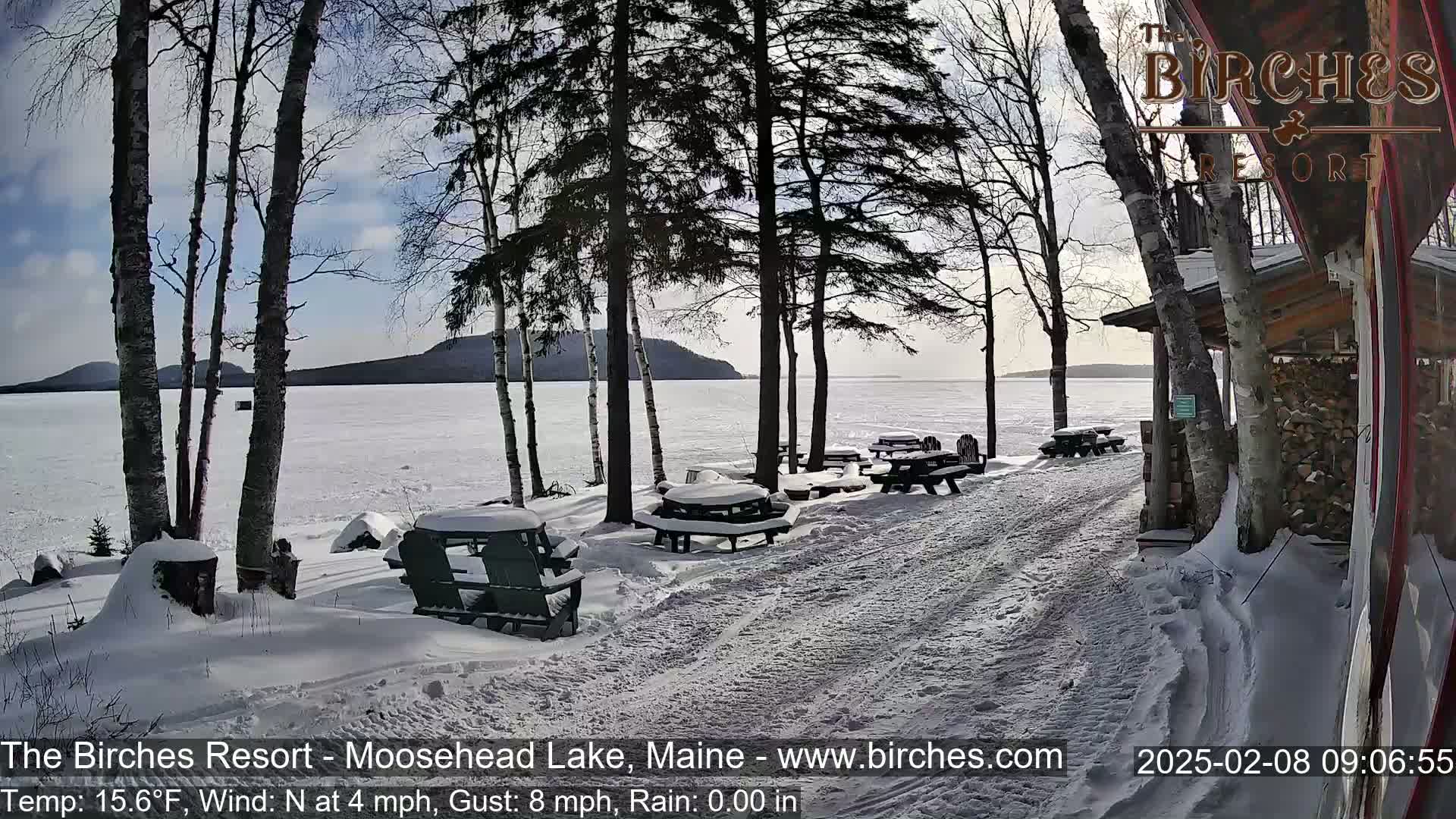 Snow-covered picnic tables and Adirondack chairs sit near a frozen lake under a partly sunny sky.