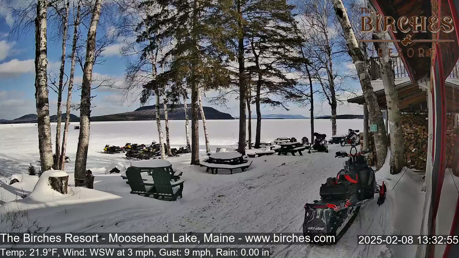 A snow-covered lakeside resort with picnic tables, snowmobiles, and birch trees under a partly sunny sky.