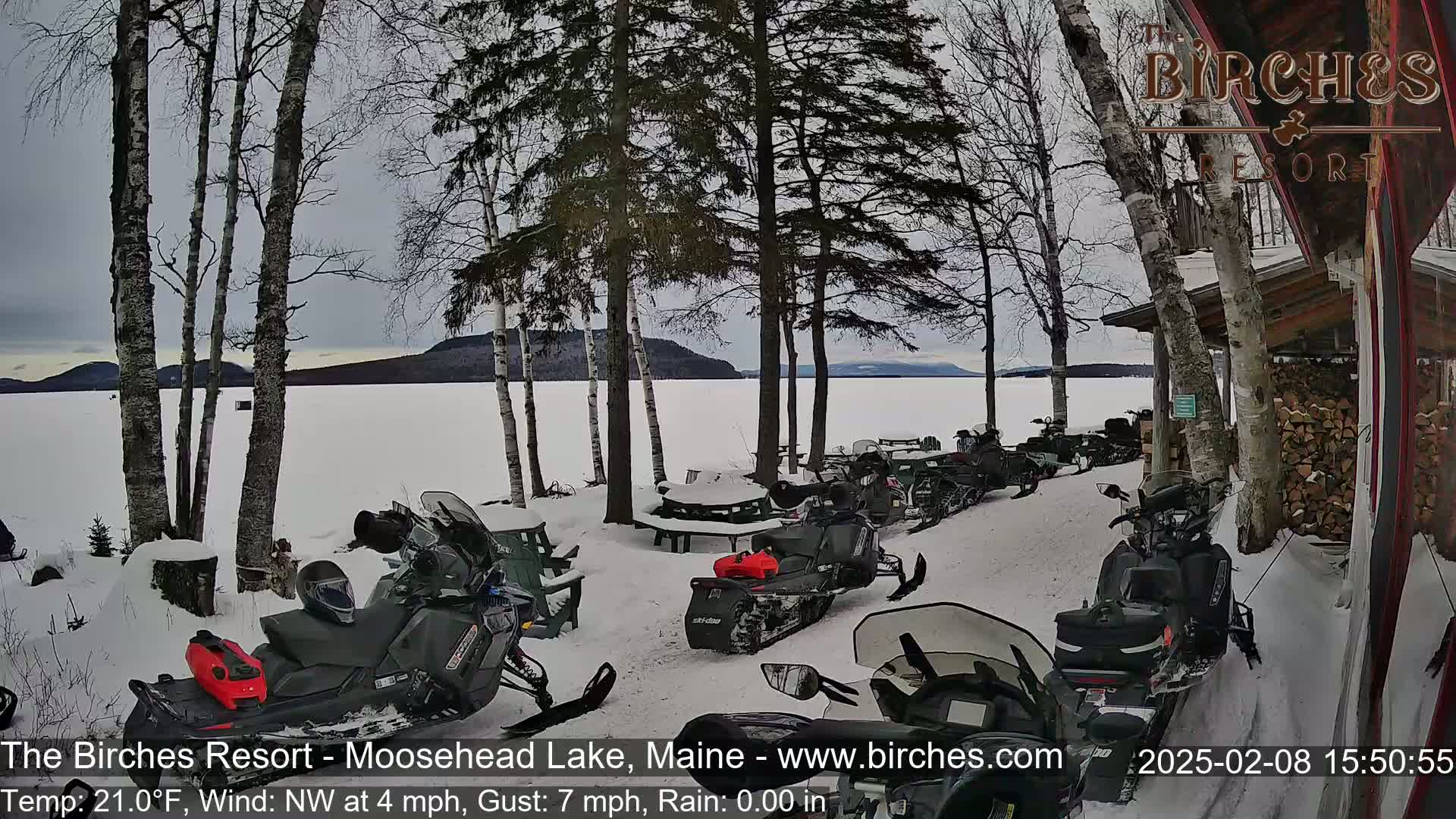 Several snowmobiles are parked near a snow-covered lakeshore, with a frozen lake and distant mountains visible under a cloudy sky.