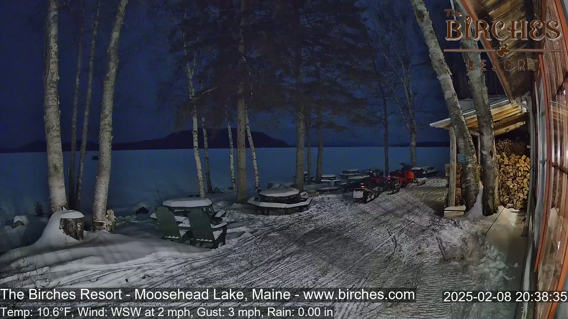 A snow-covered lakeside area at night, with several snowmobiles parked near picnic tables under birch trees, a calm lake visible in the background.