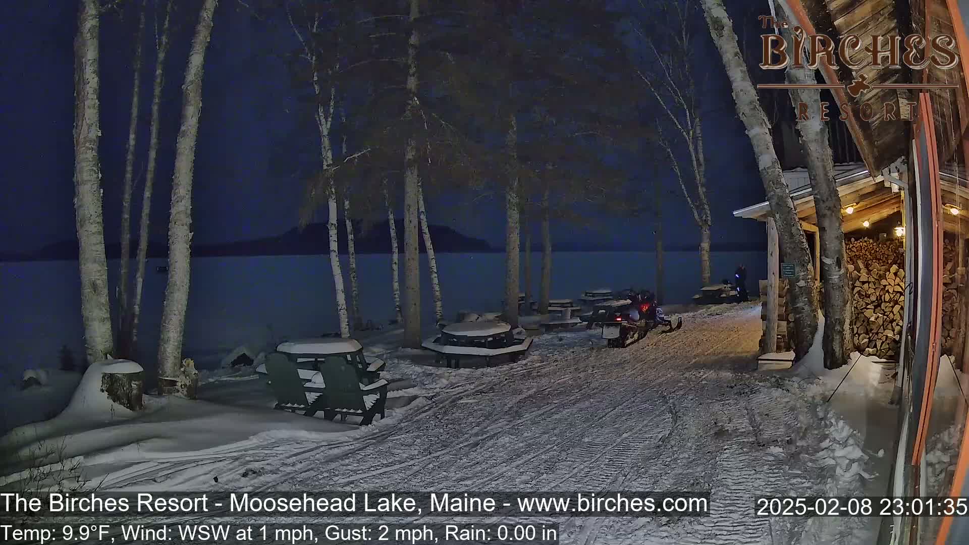 A snow-covered lakeside area at night, featuring picnic tables, Adirondack chairs, and a snowmobile near a woodpile by a building, under a dark sky with light rain.