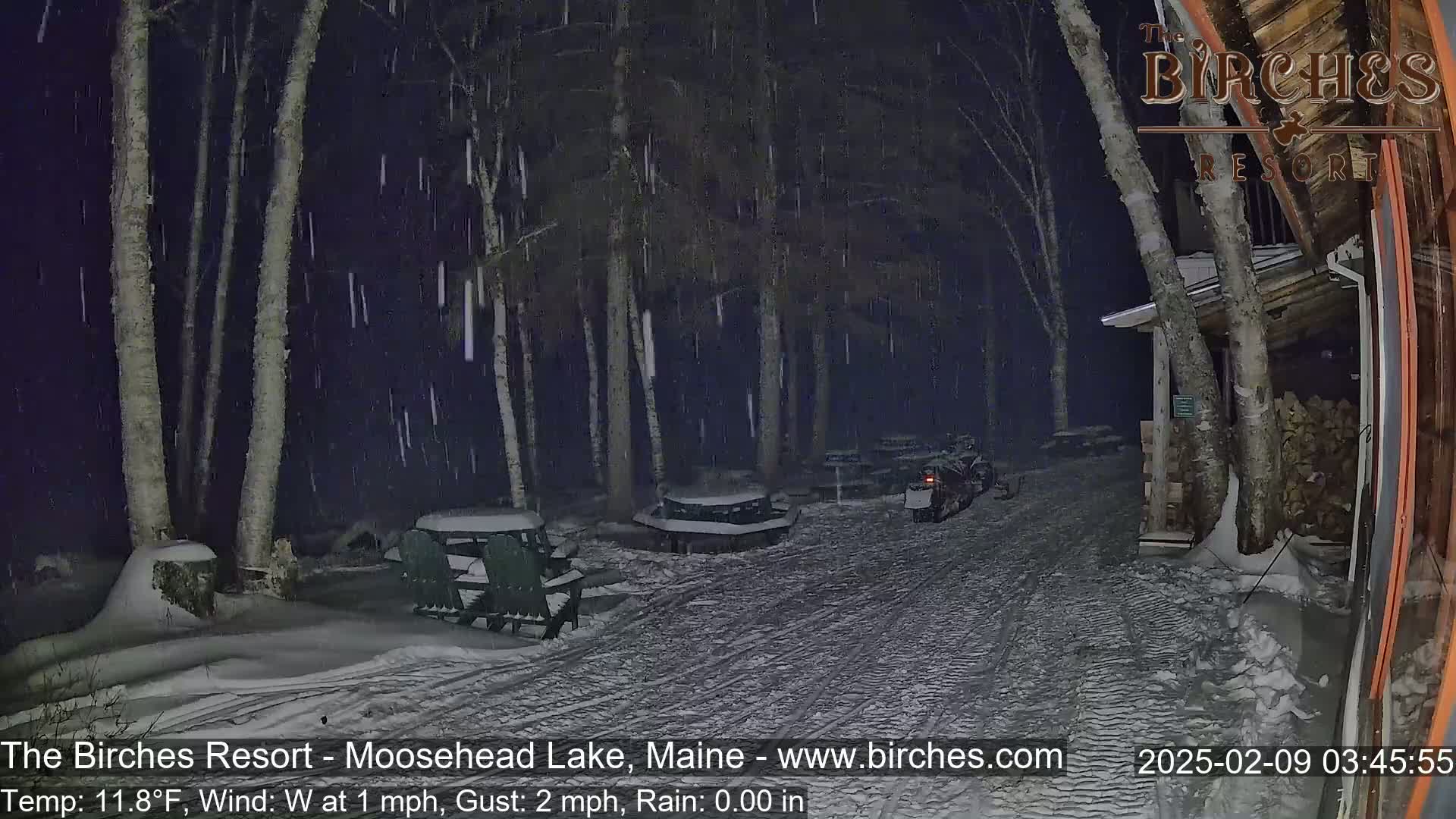 A snow-covered area with picnic tables and a snowmobile is seen at night under falling snow.