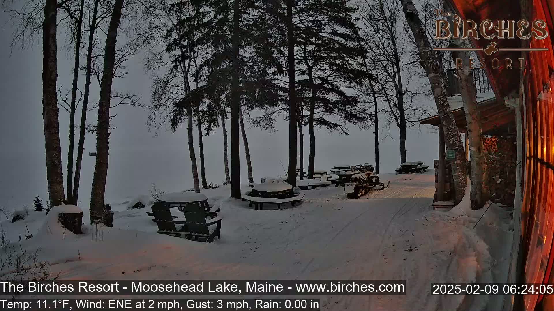 Snow-covered picnic tables and a snowmobile sit near a wooden building in a snowy, dimly lit outdoor setting.