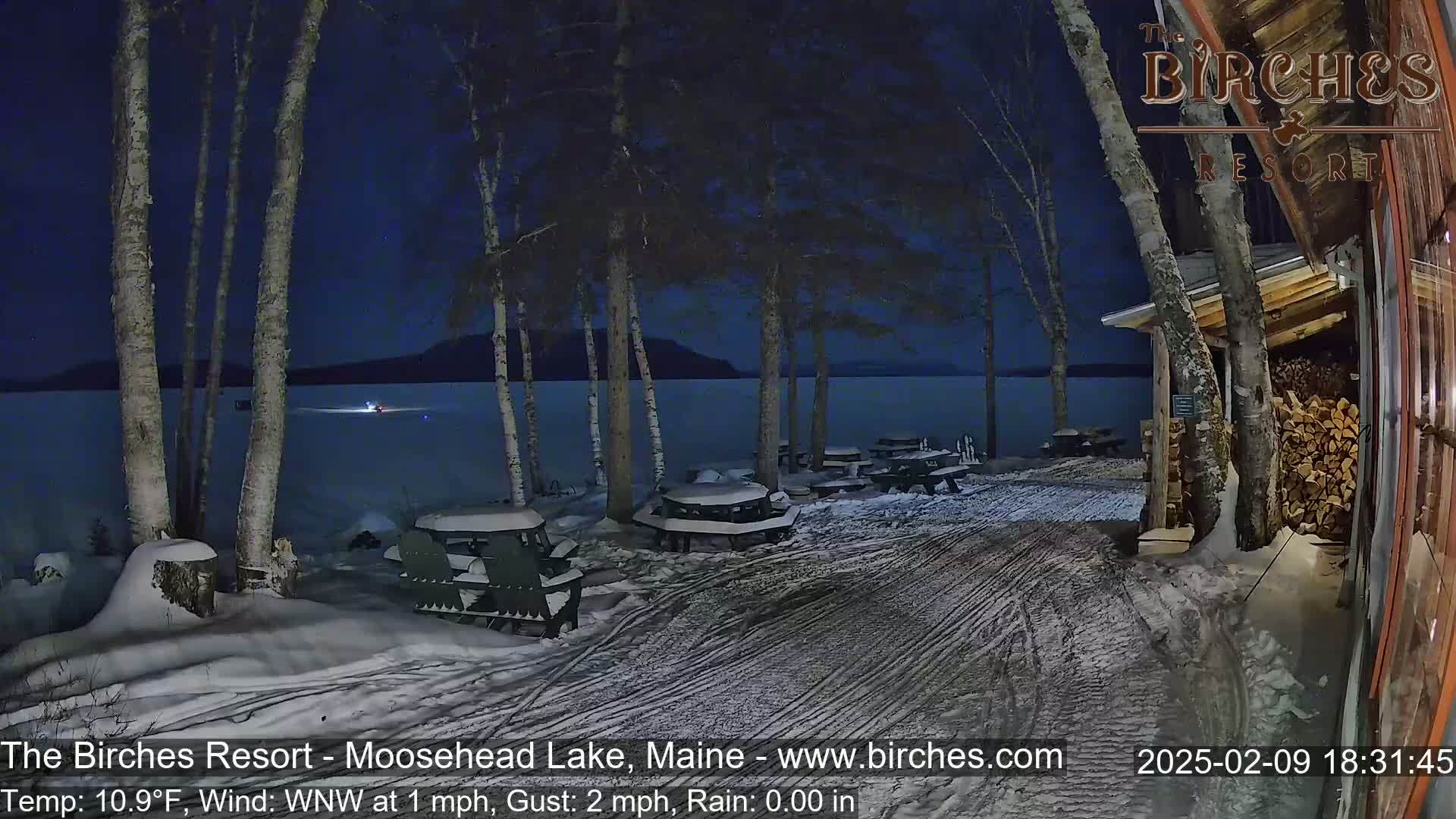 A snow-covered lakeside area at night, featuring picnic tables and Adirondack chairs near a building, under a dark blue sky.