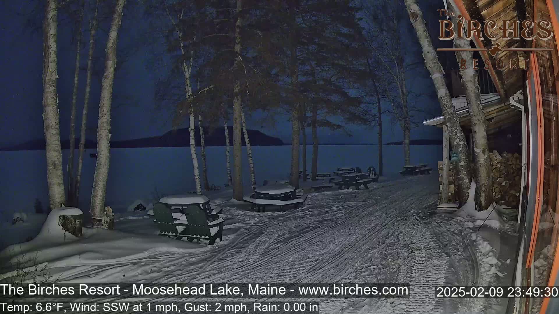 Snow-covered picnic tables and Adirondack chairs sit beside a snow-covered path leading to a lake under a dark, nighttime sky.