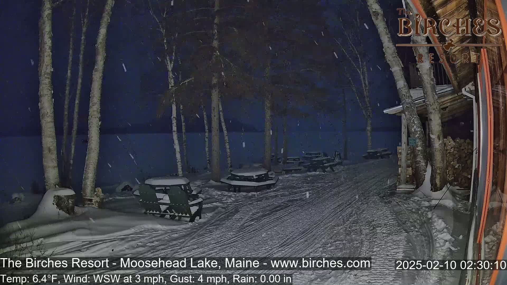 A snow-covered lakeside patio area at night, under a lightly falling snow, with picnic tables and chairs.