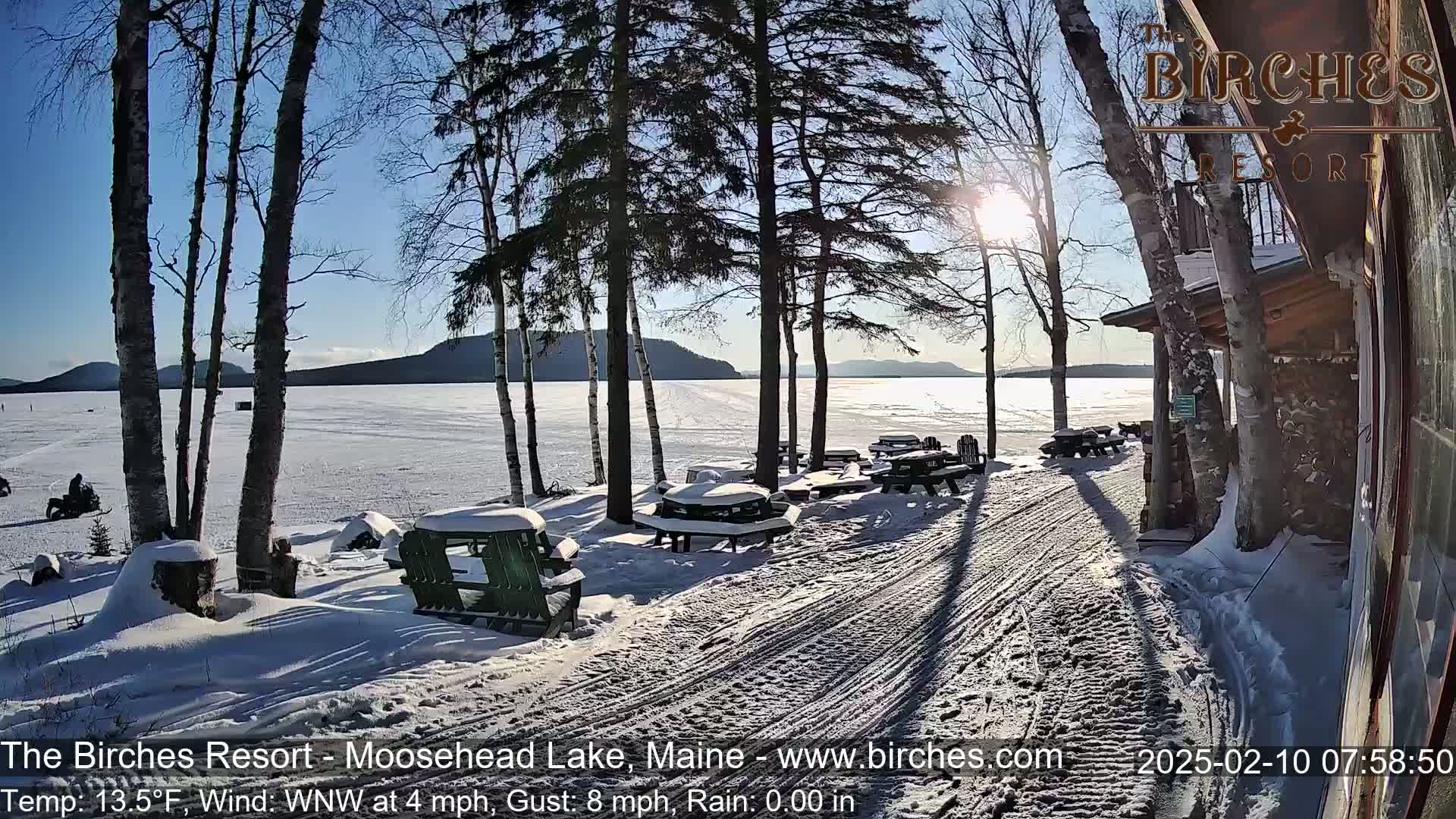 A snow-covered lakeside area with picnic tables and chairs near a building, under a sunny, cold winter sky.