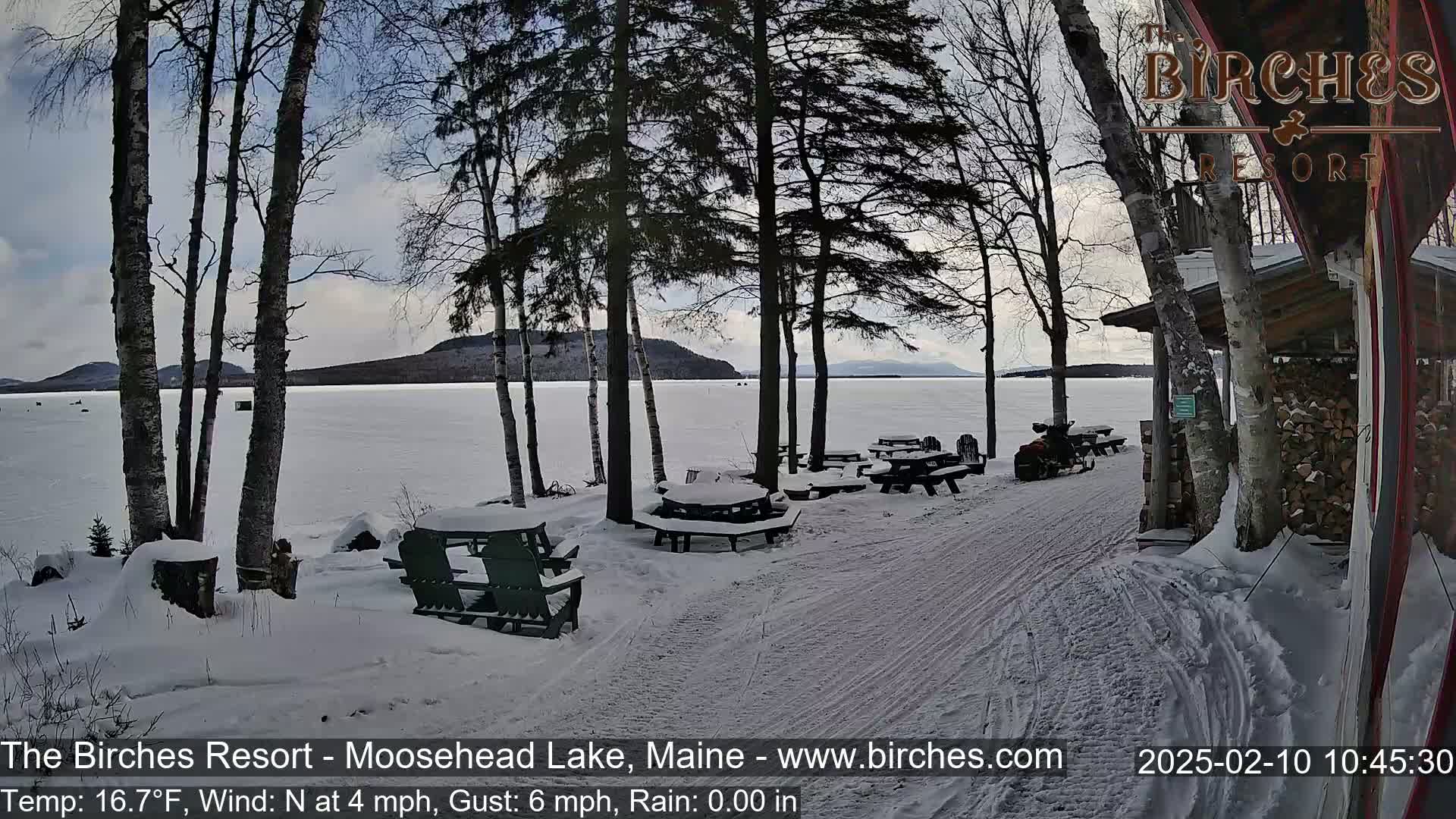Snow-covered picnic tables and chairs sit near a snow-covered shoreline with a frozen lake and distant hills under a cloudy sky.