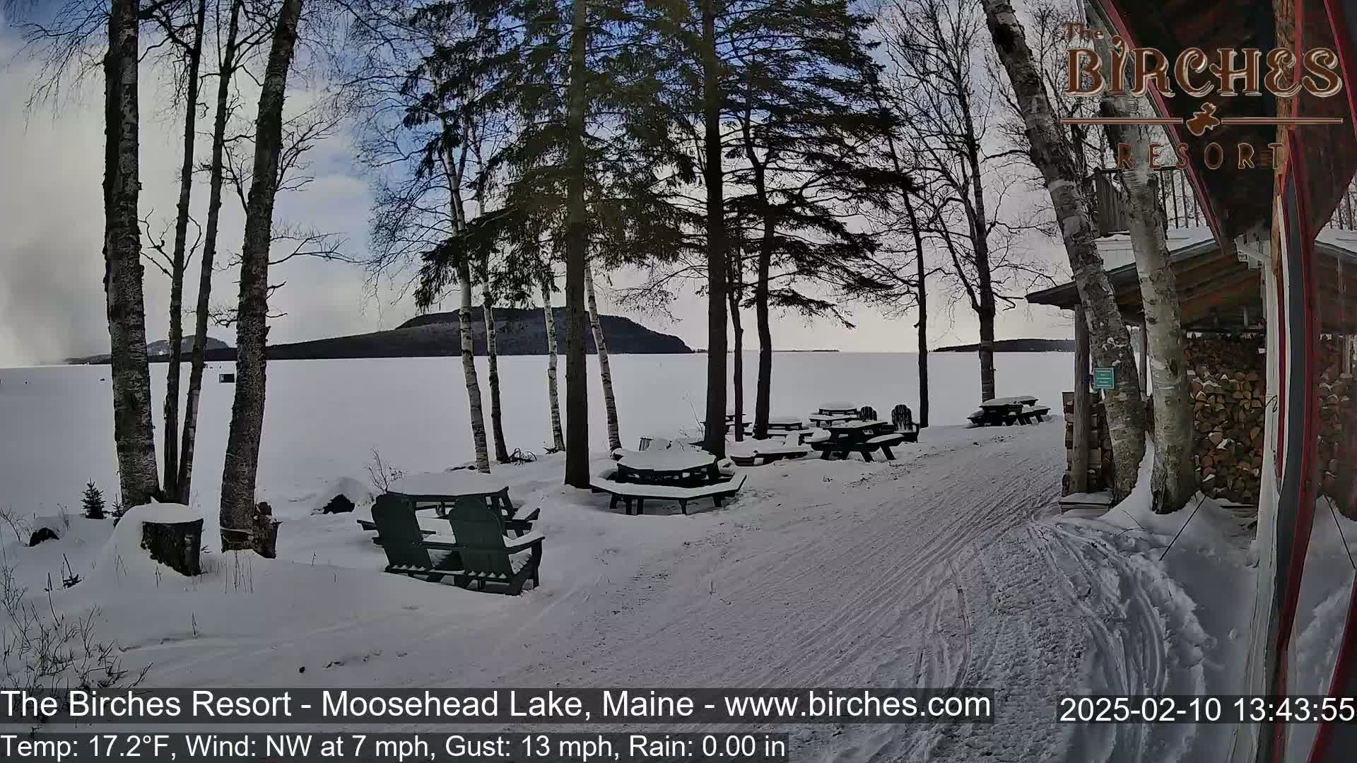 A snow-covered lakeside picnic area with several empty tables and chairs is visible, with a calm lake and distant land in the background under a partly cloudy sky.