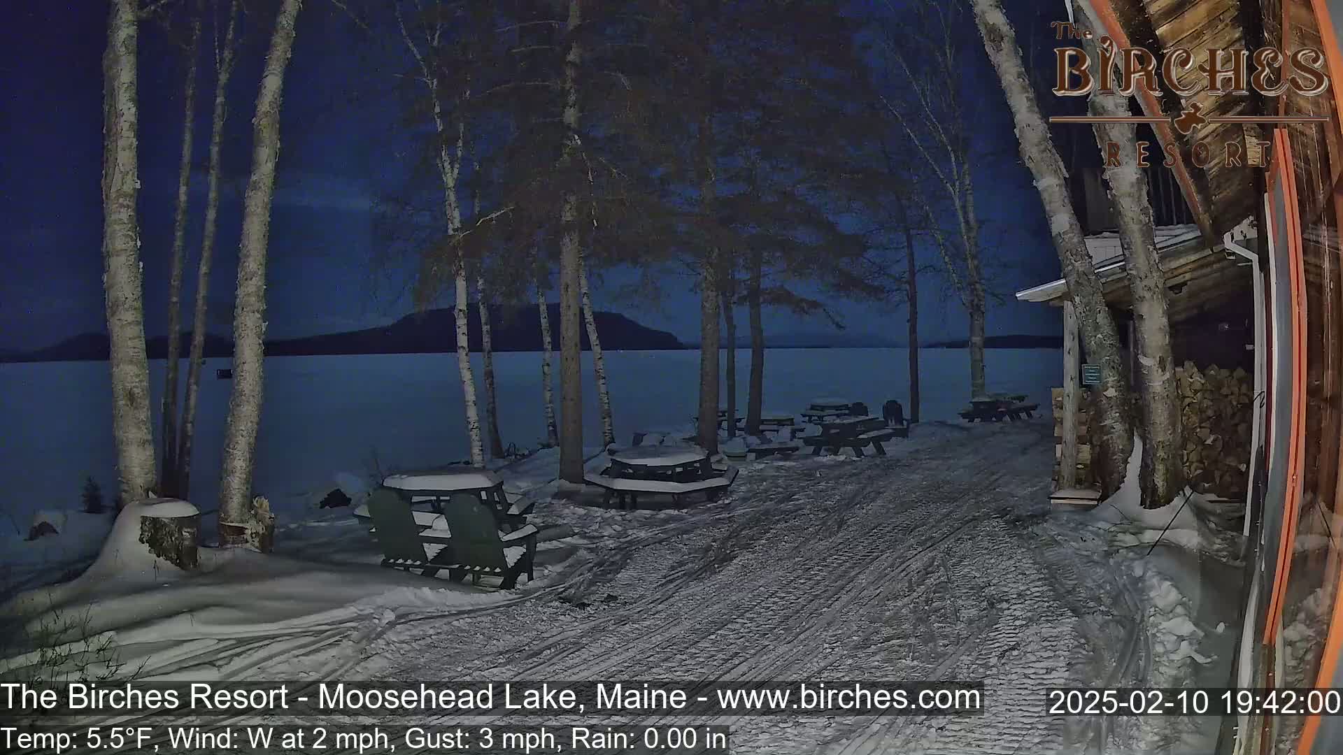 A snow-covered lakeside patio with picnic tables and chairs is visible at night under a dark blue sky, with a calm lake and distant landmass in the background.
