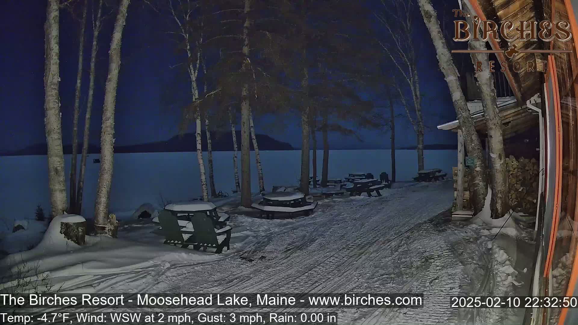 A snow-covered lakeside patio area at night, under a dark blue sky, with several snow-covered picnic tables and chairs near a building.