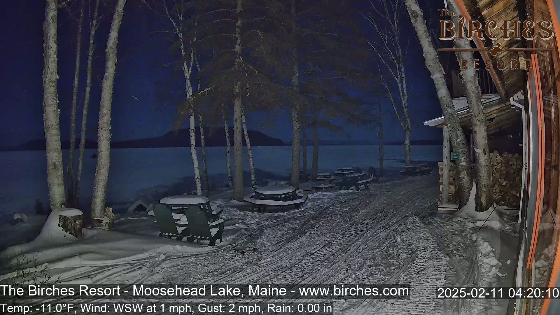 A snow-covered lakeside area at night shows several picnic tables and chairs under birch trees near a building, with a calm lake visible in the background under a dark blue sky.