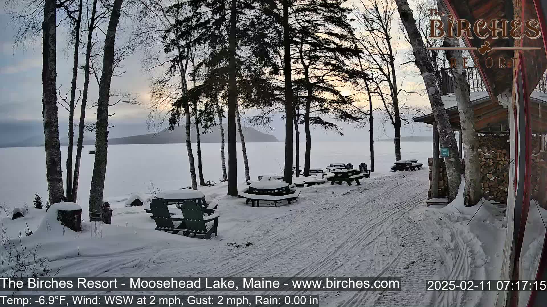 A snow-covered lakeside picnic area with several empty tables and chairs, situated near a building with a woodpile, under a partly cloudy sky.