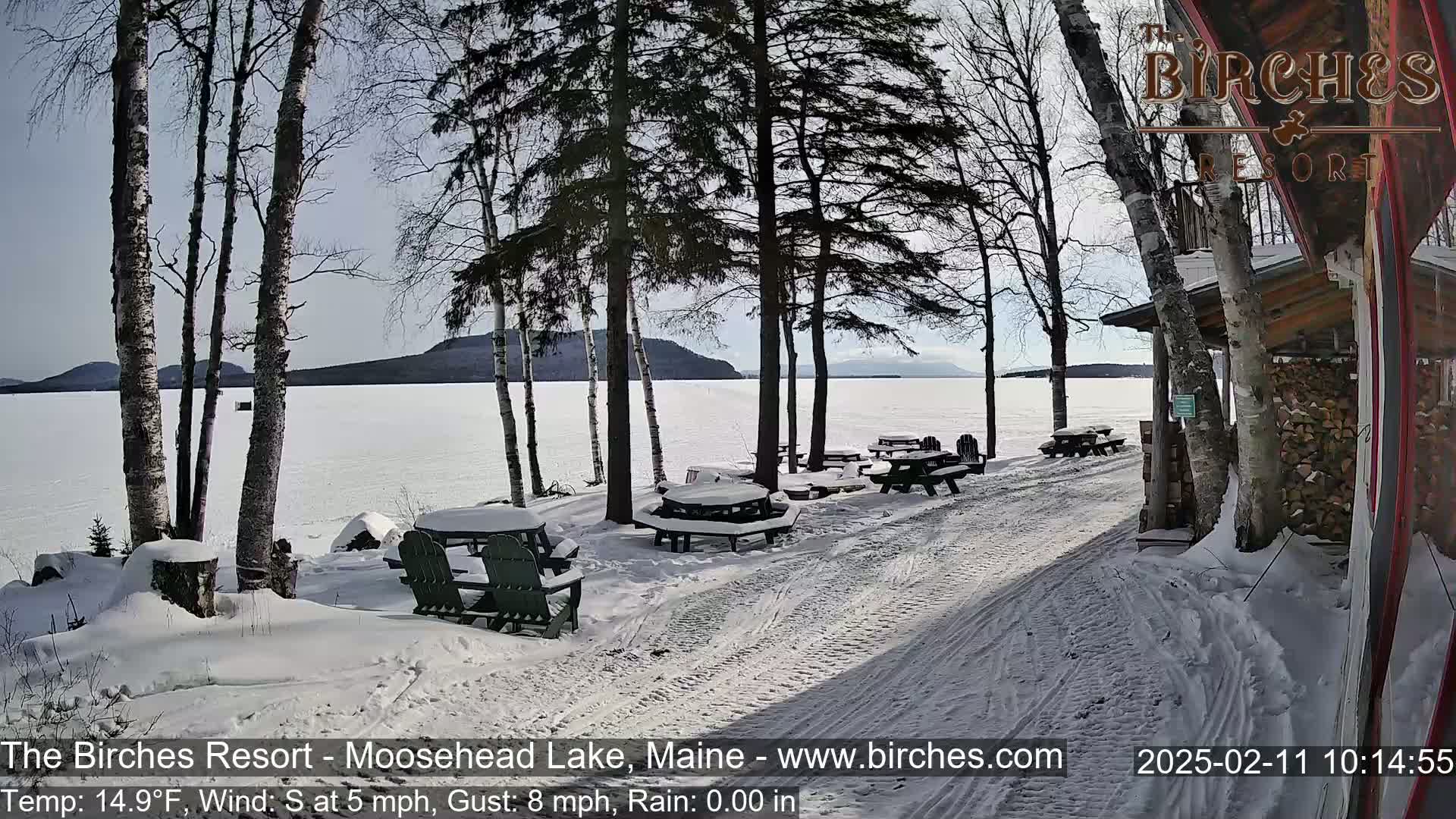 Snow-covered picnic tables and chairs sit near a frozen lake under a sunny sky, with bare trees and a wooden building visible.