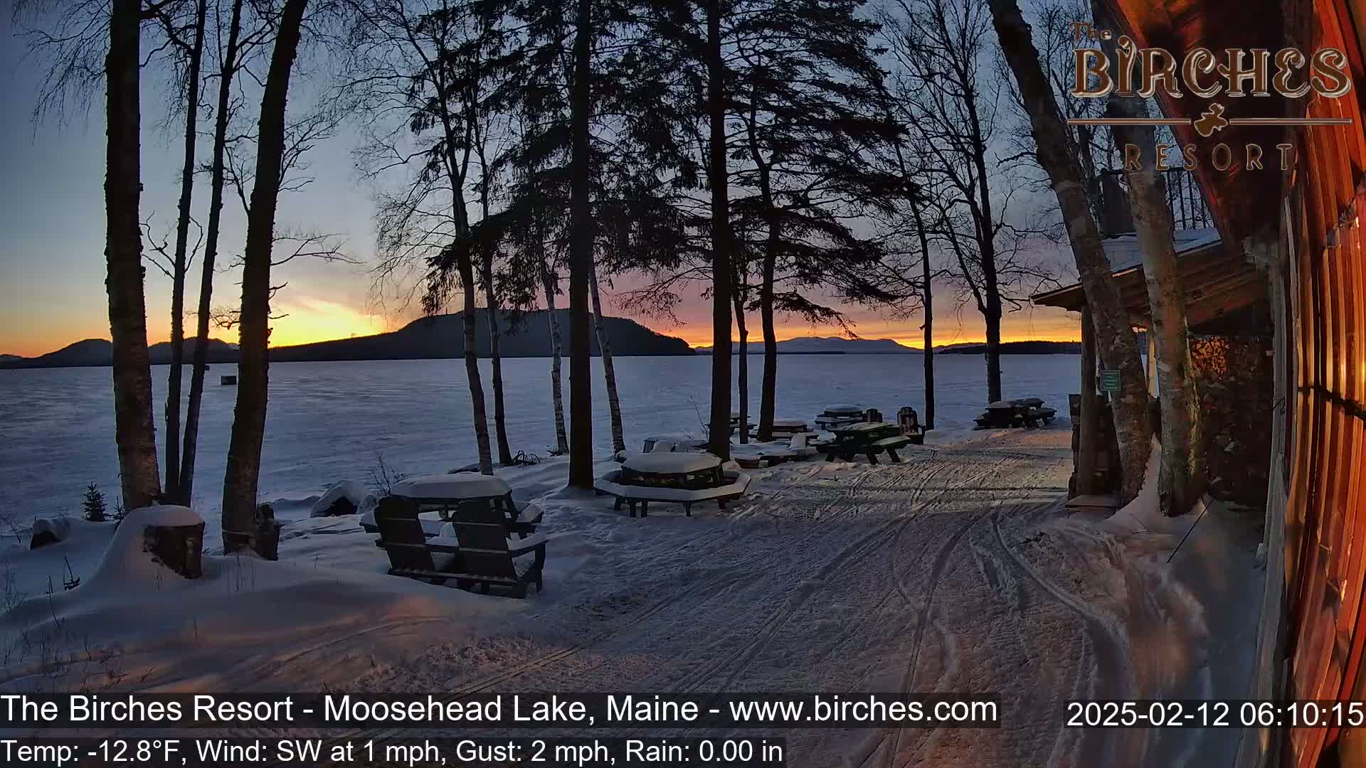 A snowy lakeside scene at sunset, with several snow-covered picnic tables and chairs, and birch trees silhouetted against a colorful sky.