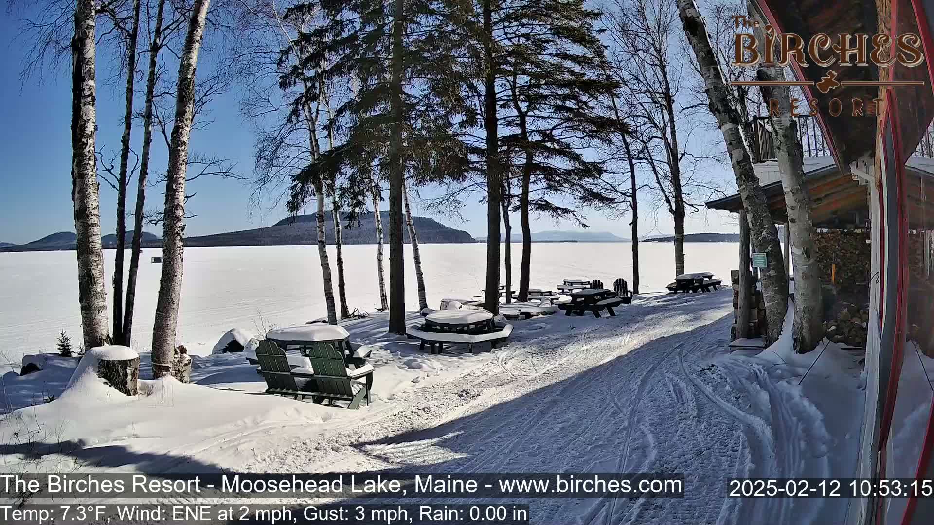 A snow-covered lakeside scene with picnic tables and Adirondack chairs under a sunny sky.