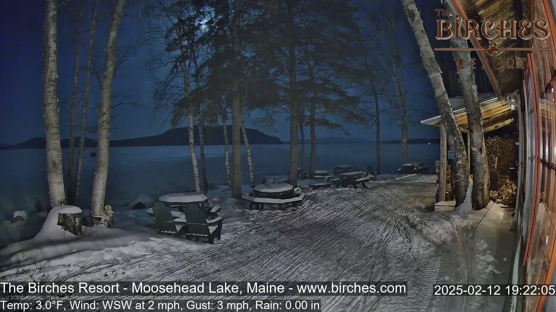 A snow-covered lakeside area at night, under a partly cloudy moonlit sky, features several snow-dusted picnic tables and chairs near a building with birch trees in the foreground.