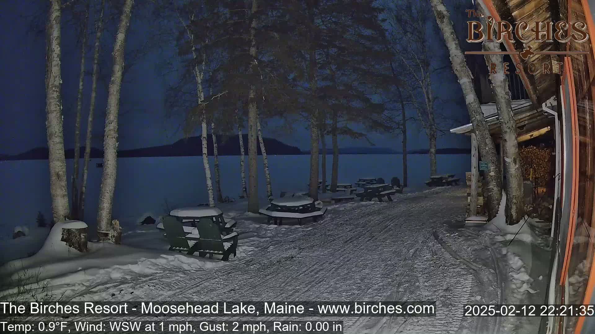 Snow-covered picnic tables sit near a snow-covered lakeshore at night under a dark sky.