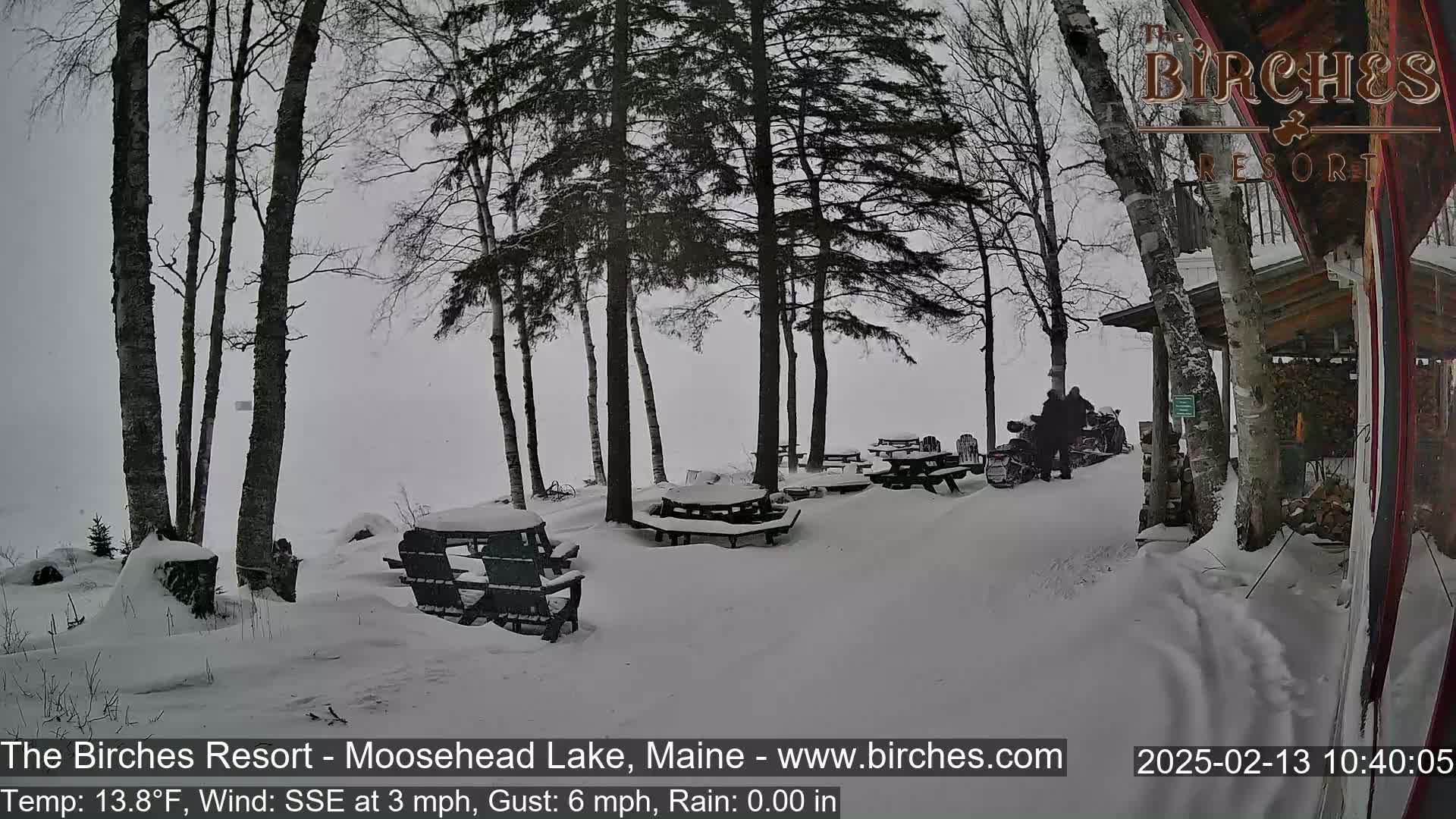 Snow-covered picnic tables and chairs sit beneath snow-laden trees in a snowy, overcast landscape, with a snowmobile parked nearby.