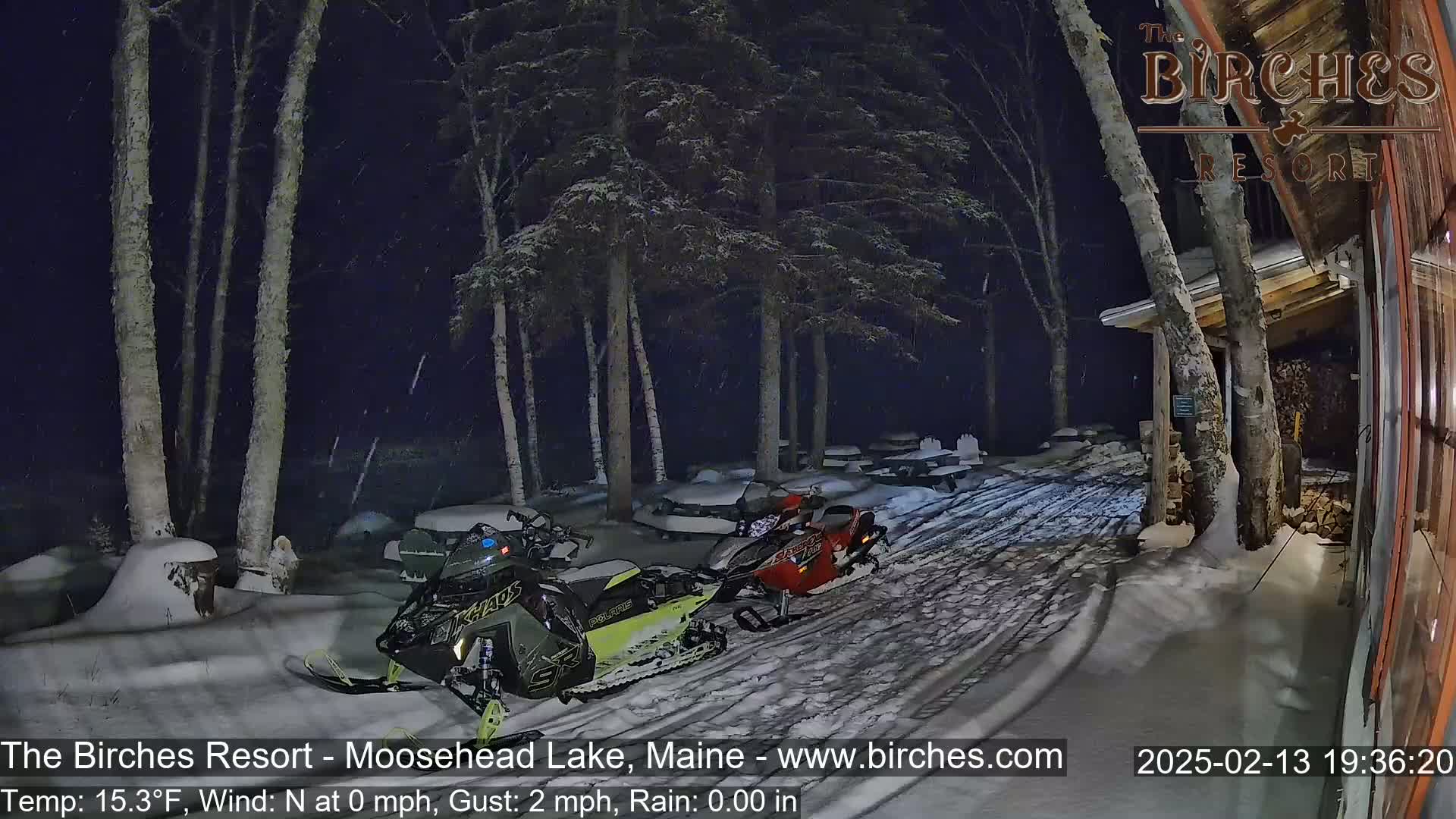 Two snowmobiles are parked in a snow-covered area near a building at night, under light snowfall.