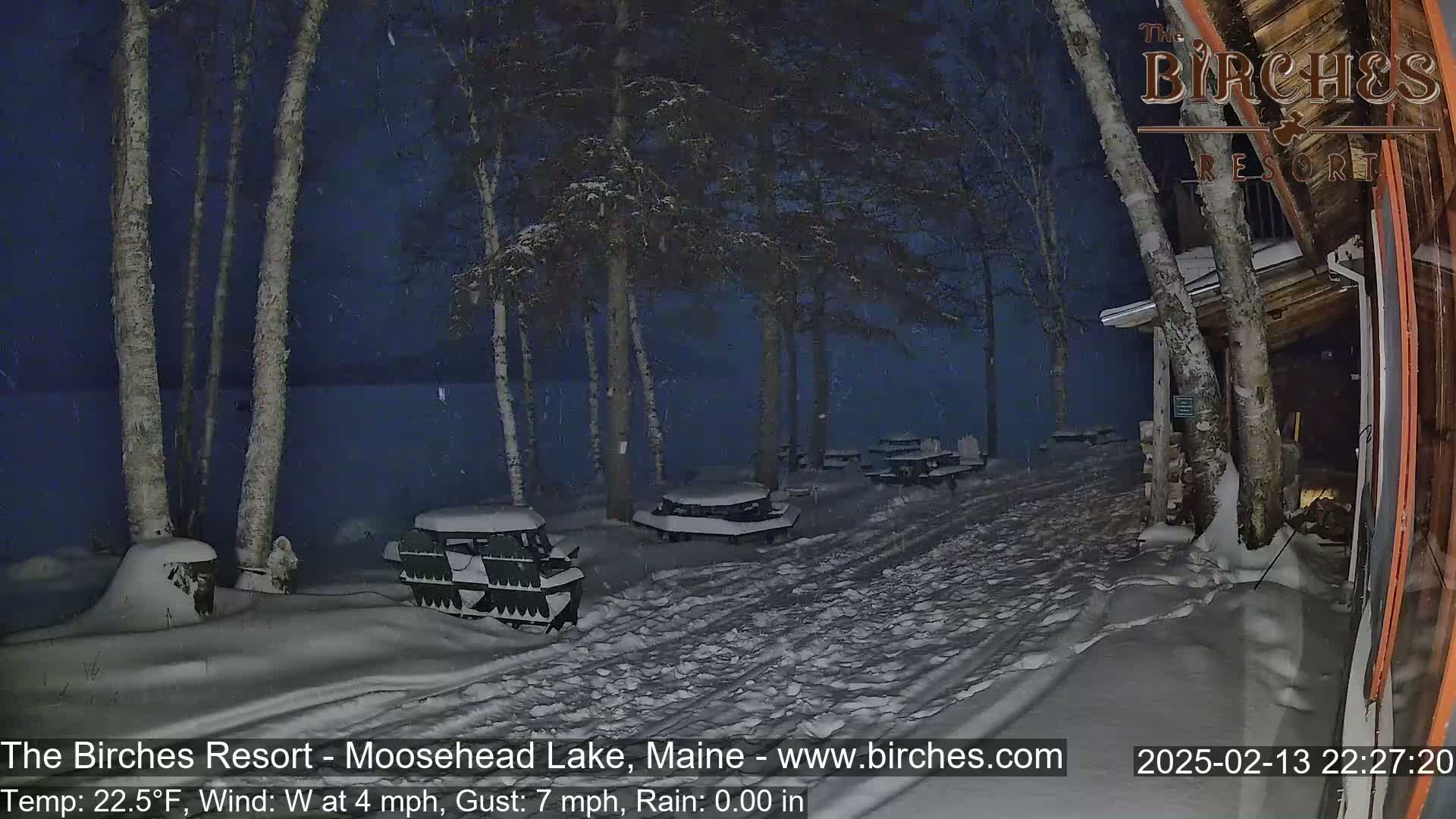 Snow falls at night on a snow-covered path leading to several snow-covered picnic tables near a lake and a building.