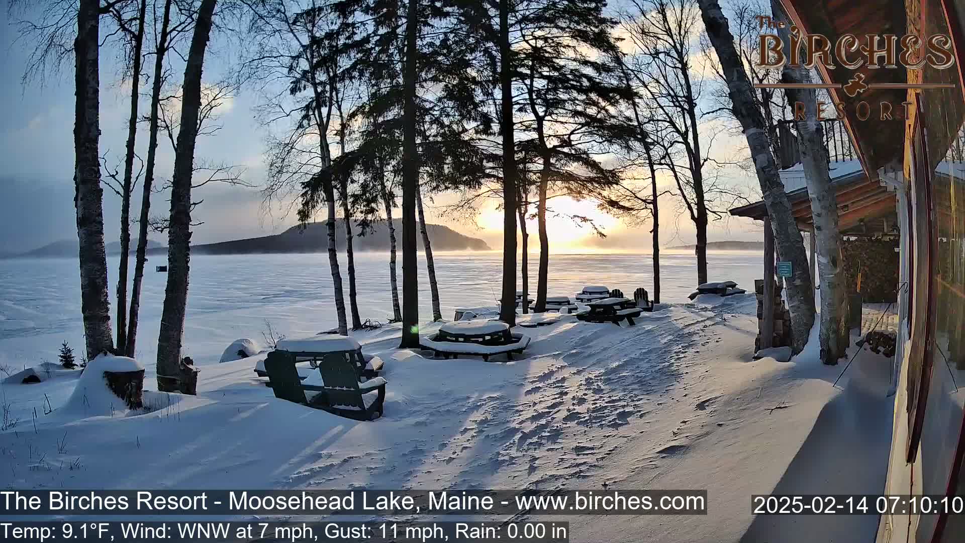 A snow-covered lakeside scene at sunrise shows several snow-covered picnic tables and chairs near a building, with trees in the foreground and a frozen lake beyond.