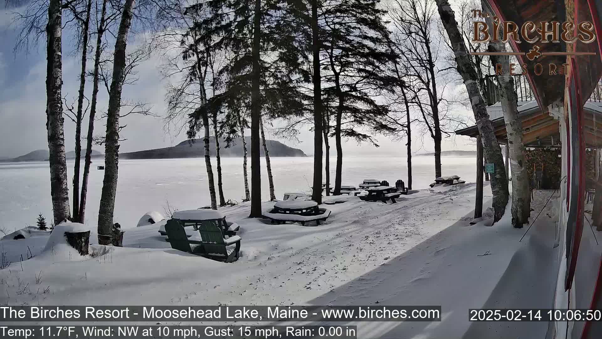 A snow-covered lakeside patio with picnic tables and chairs sits under a partly cloudy sky, with a calm frozen lake and distant land in the background.